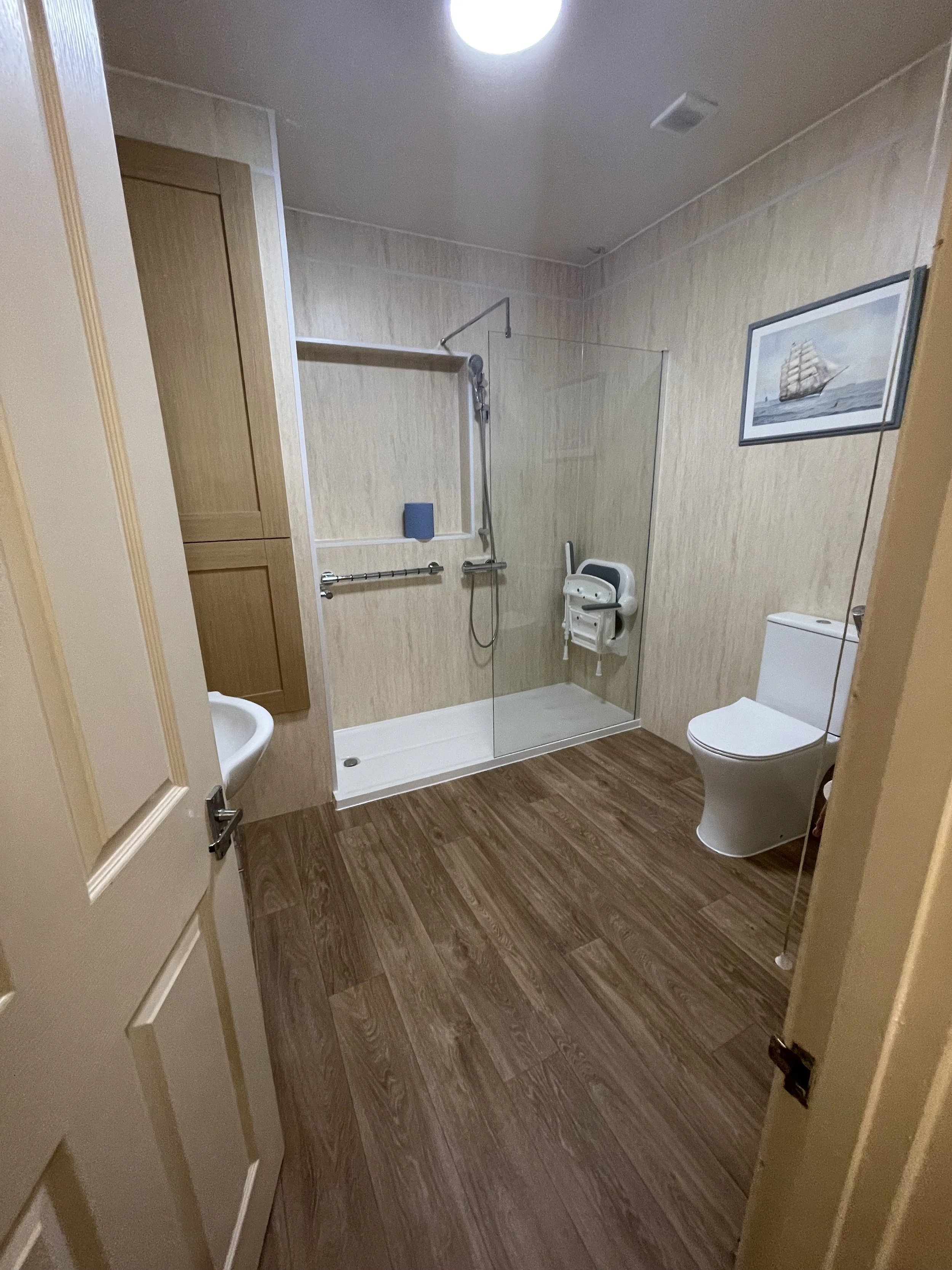 Bathroom with wooden flooring, walk-in shower with glass door, toilet, small sink, wall artwork of a sailing ship, and a wall-mounted shower chair.