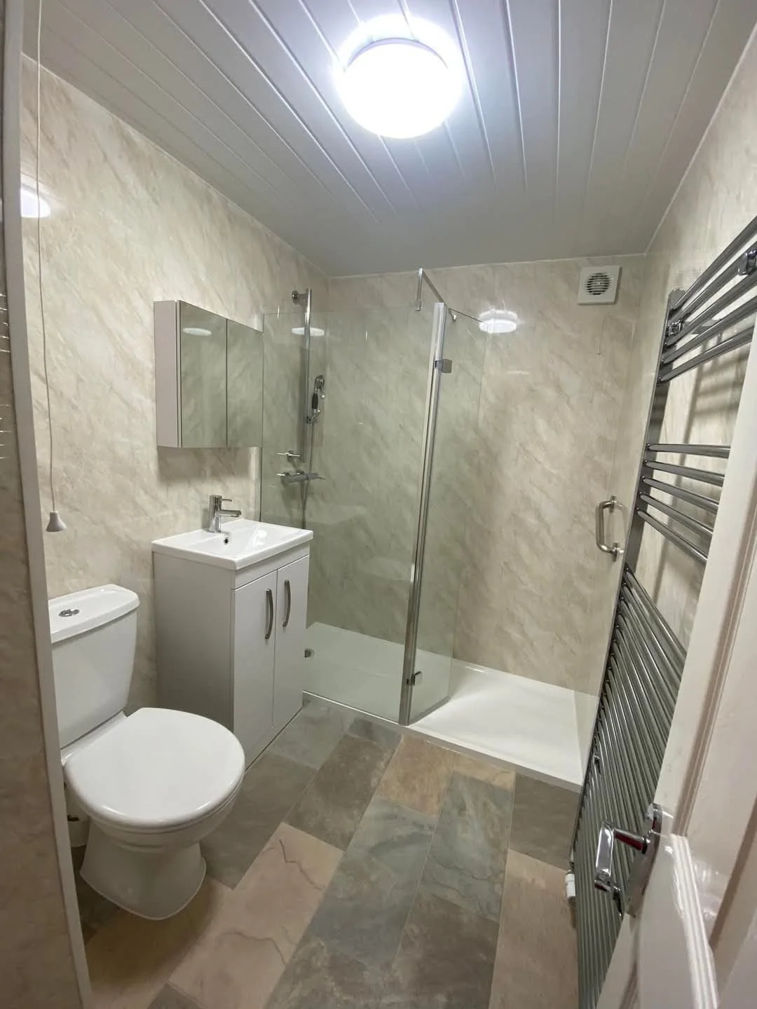 Small bathroom with a toilet, a white vanity with a sink, a mirrored cabinet, a glass-enclosed shower, and a heated towel rail on cream-colored tiled walls and a multi-colored tiled floor.