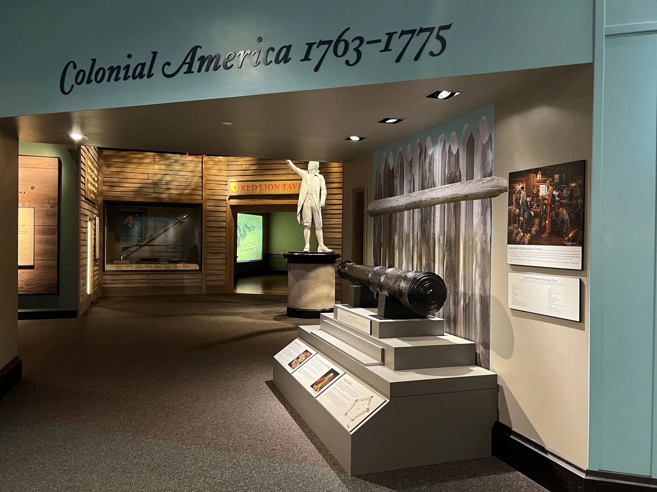 Interior of a history museum with a sign reading 'Colonial America 1763-1775'. Display of a large, old cannon, a statue of a man in colonial attire, and framed historical images and information panels.
