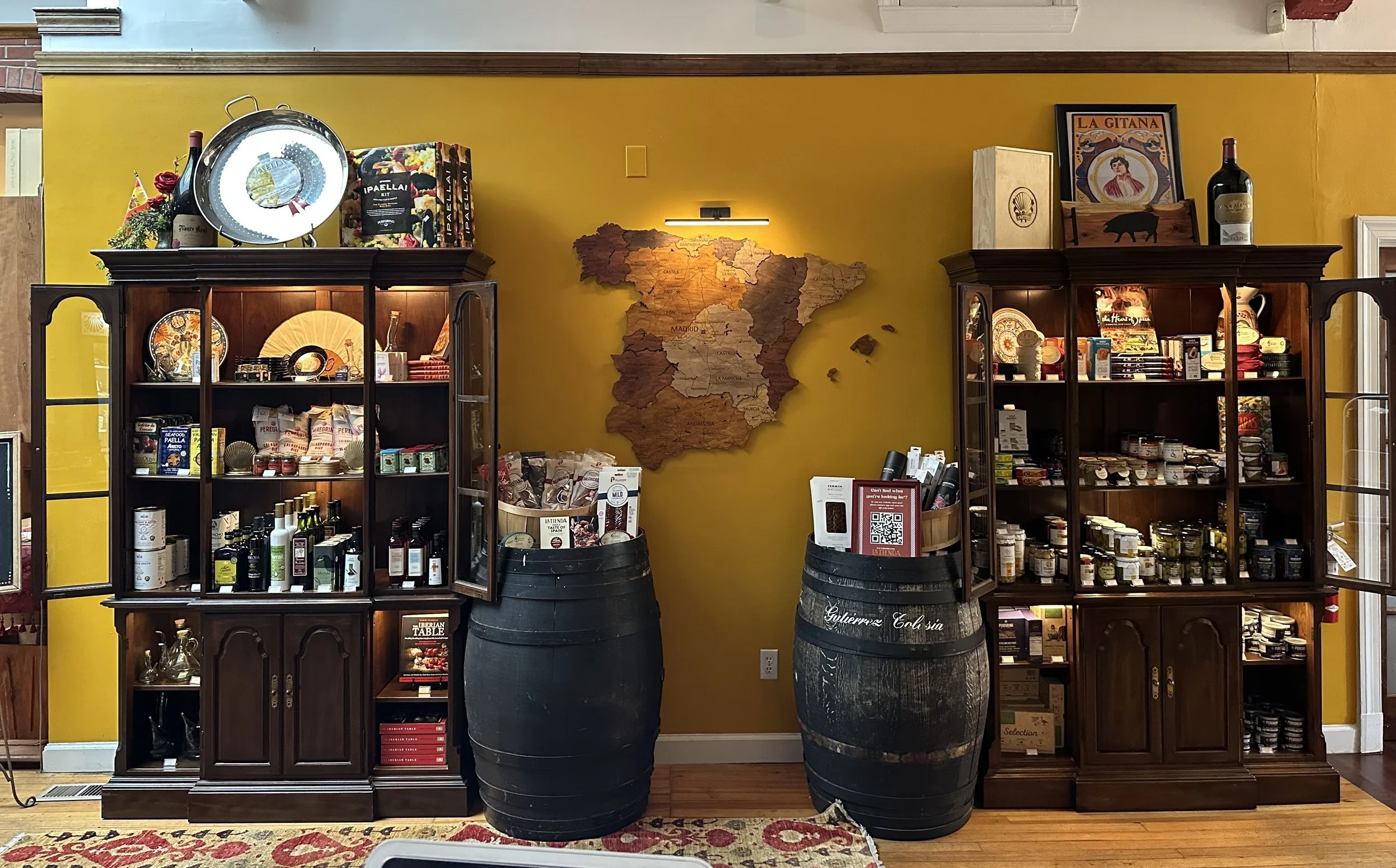 Two wooden display cabinets filled with bottles, jars, and packaged foods in a room with a yellow wall. Two large black barrels in front of the wall hold brochures and QR codes. A map of Spain is mounted on the wall, with various items and framed art displayed on top of the cabinets.