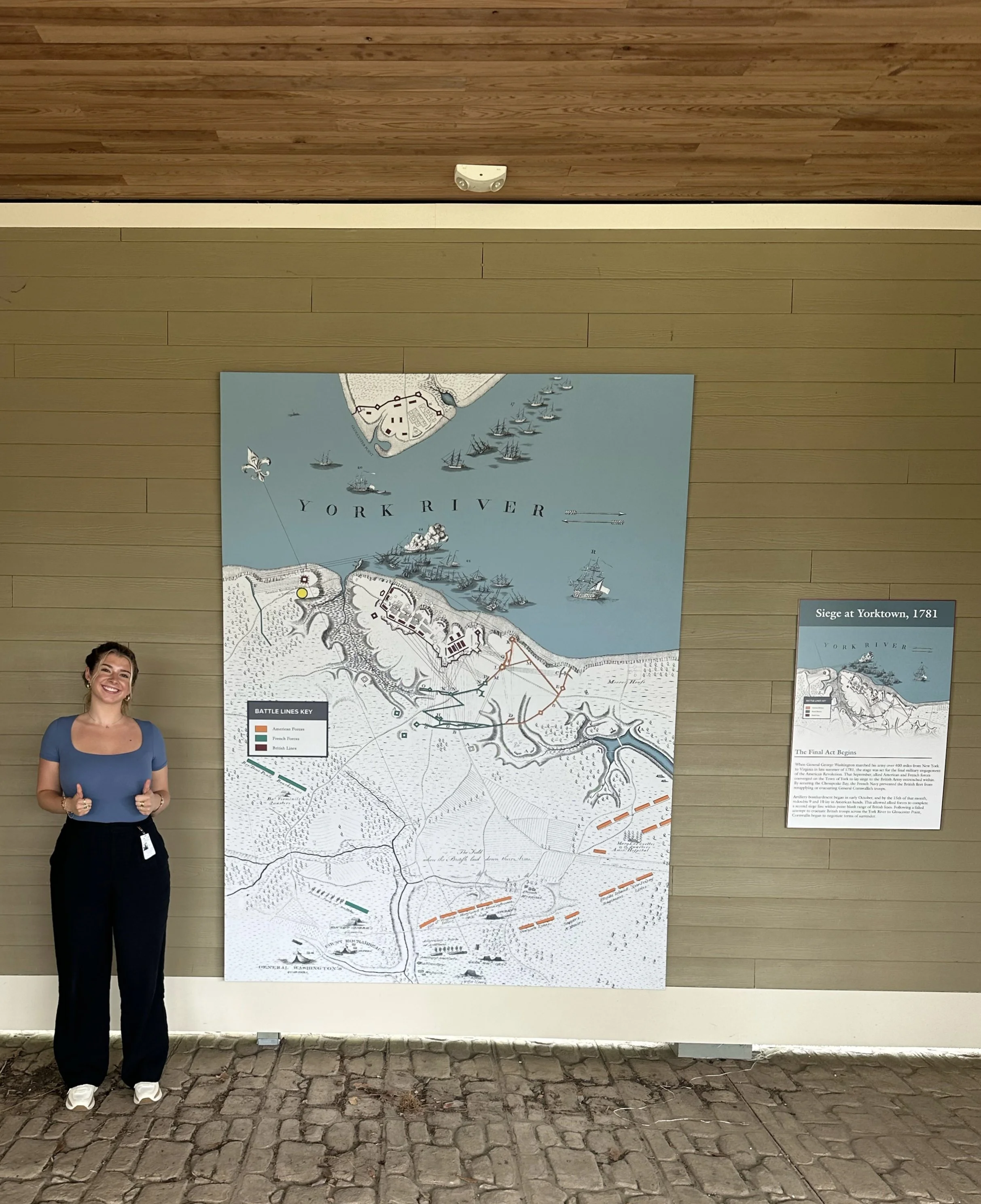Clara Fernandez standing next to a historical map of York River and Yorktown, with a smaller informational panel beside it, both mounted on a wood-paneled wall.