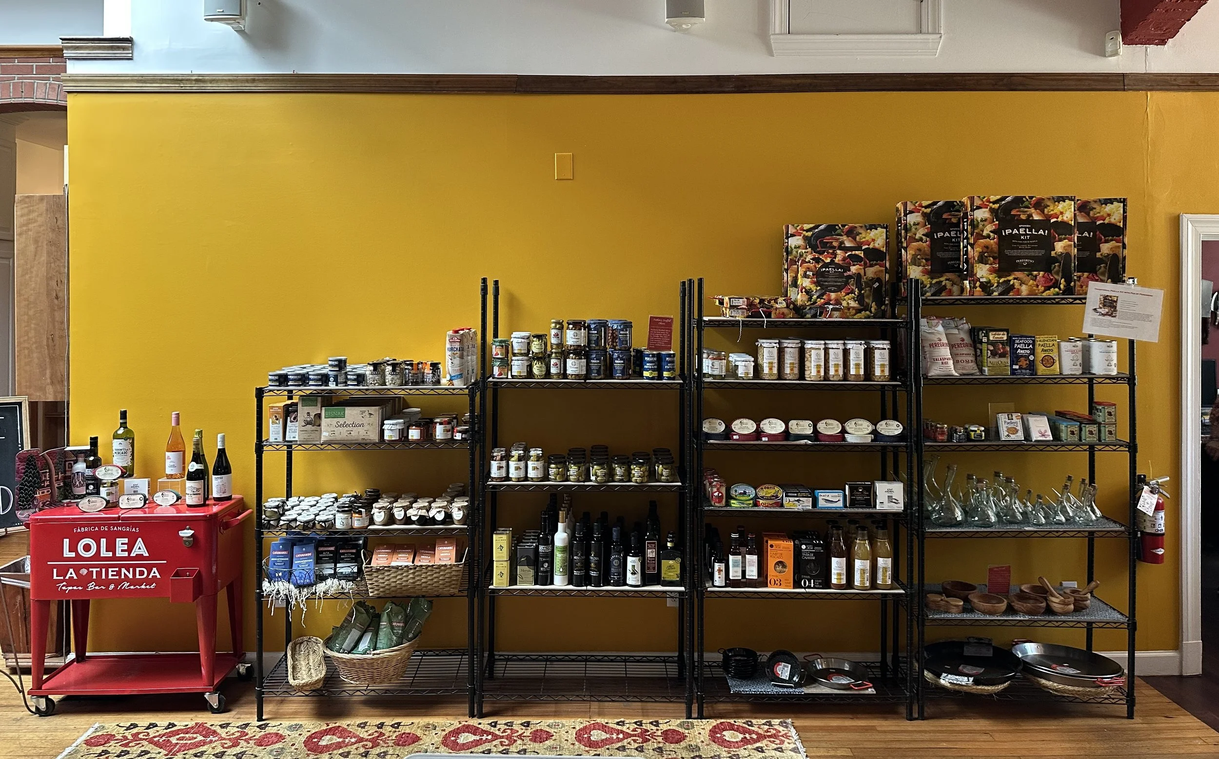 Retail store display with jars, bottles, and kitchenware against a yellow wall, including wine bottles, spices, cooking oils, and decorative bowls.