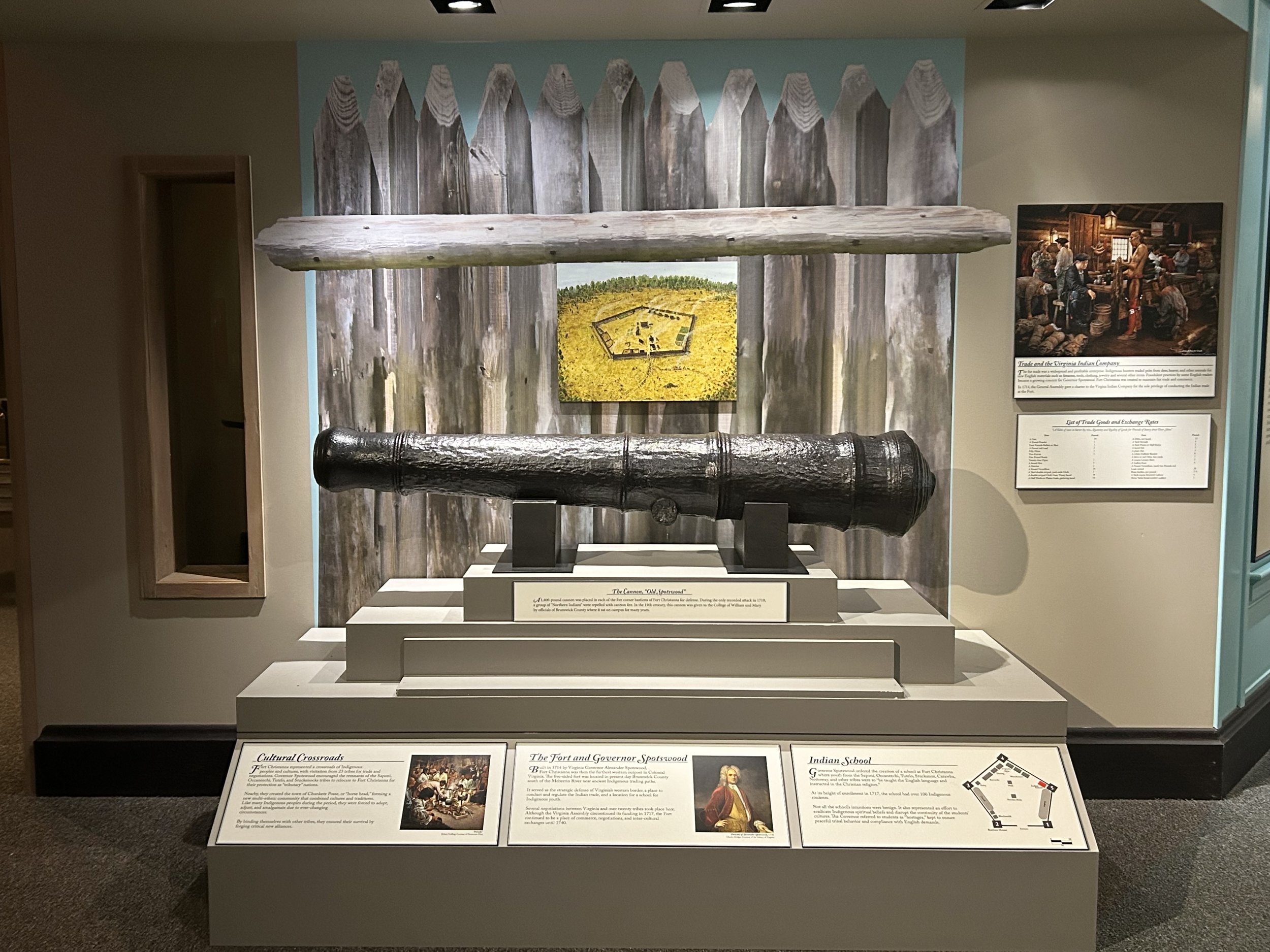 Historical cannon displayed in a museum exhibit with informational panels and a wooden fence backdrop.