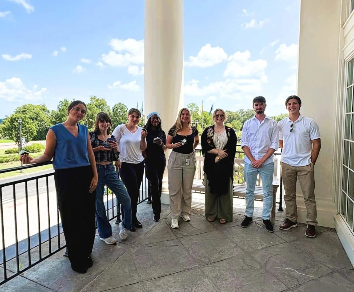 Group of nine college-aged people standing on a porch outdoors, smiling, with a view of trees and blue sky in the background.