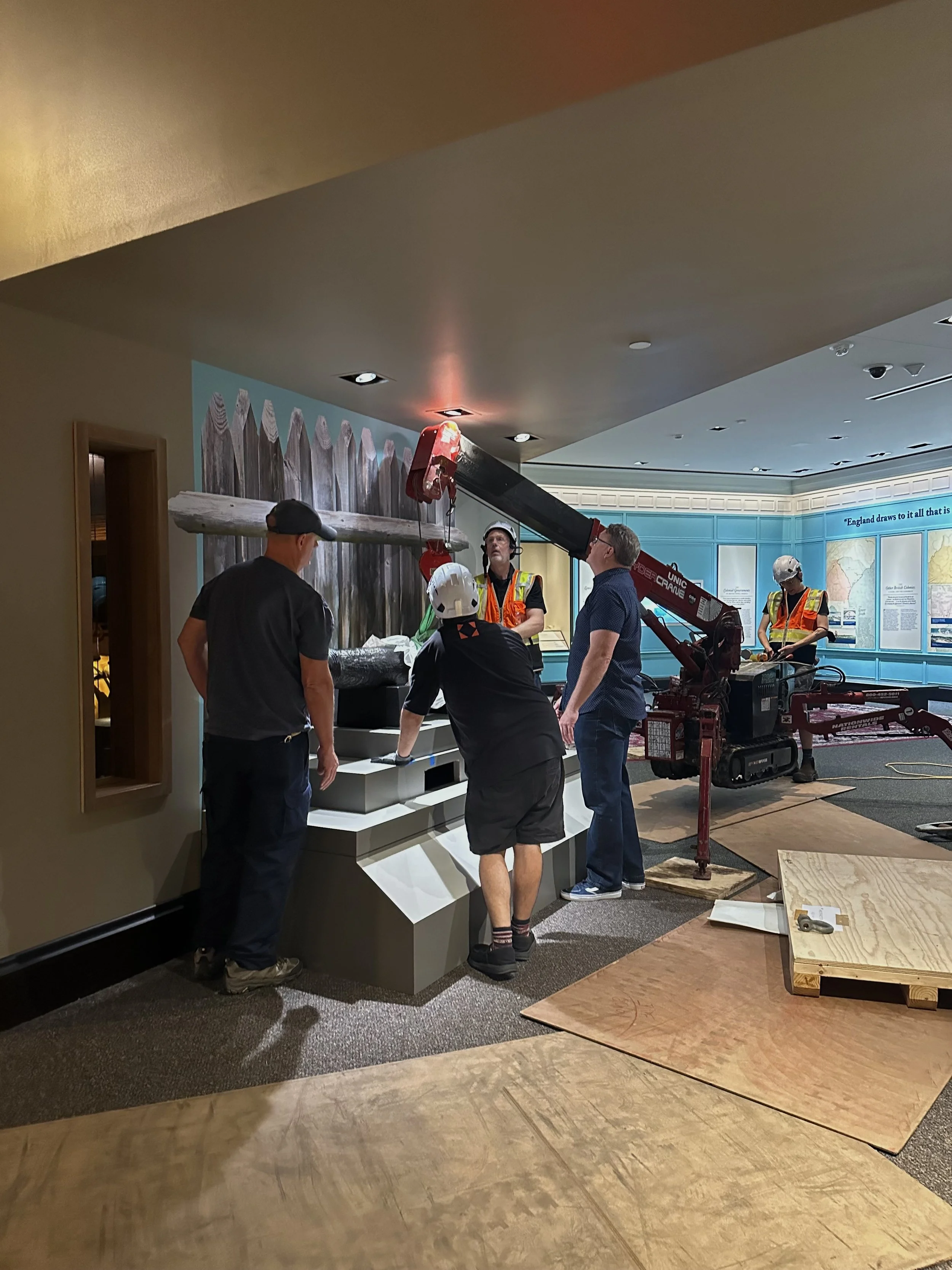 Construction workers installing a display of a large wooden fence with a log lying on top in an indoor exhibition space.