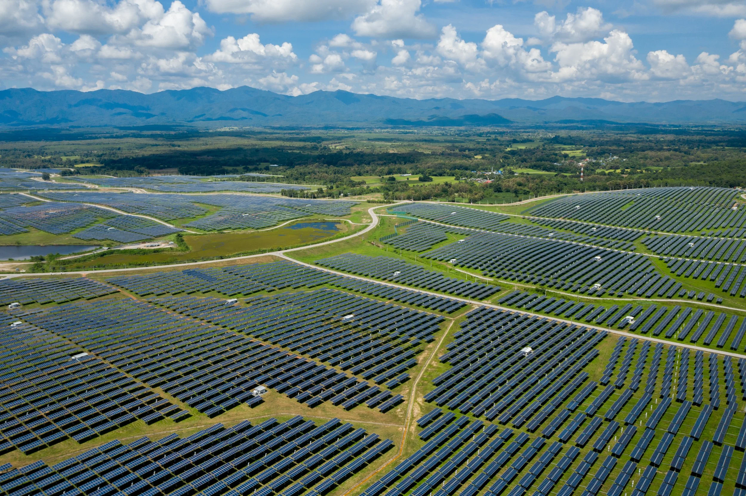 Luftaufnahme von großen Solarzellenfeldern in einer ländlichen Landschaft mit Bergen im Hintergrund unter einem bewölkten Himmel.