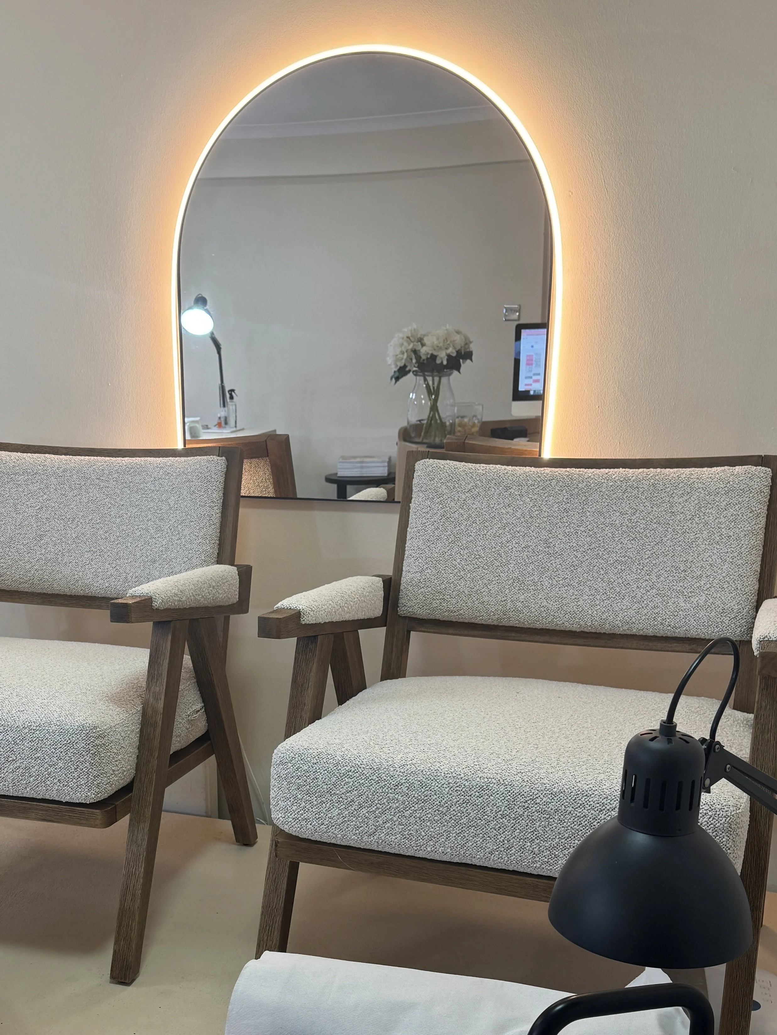 Modern seating area with two wooden chairs featuring light upholstery, an arched backlit mirror on the wall, a small side table with flowers, and a black desk lamp.