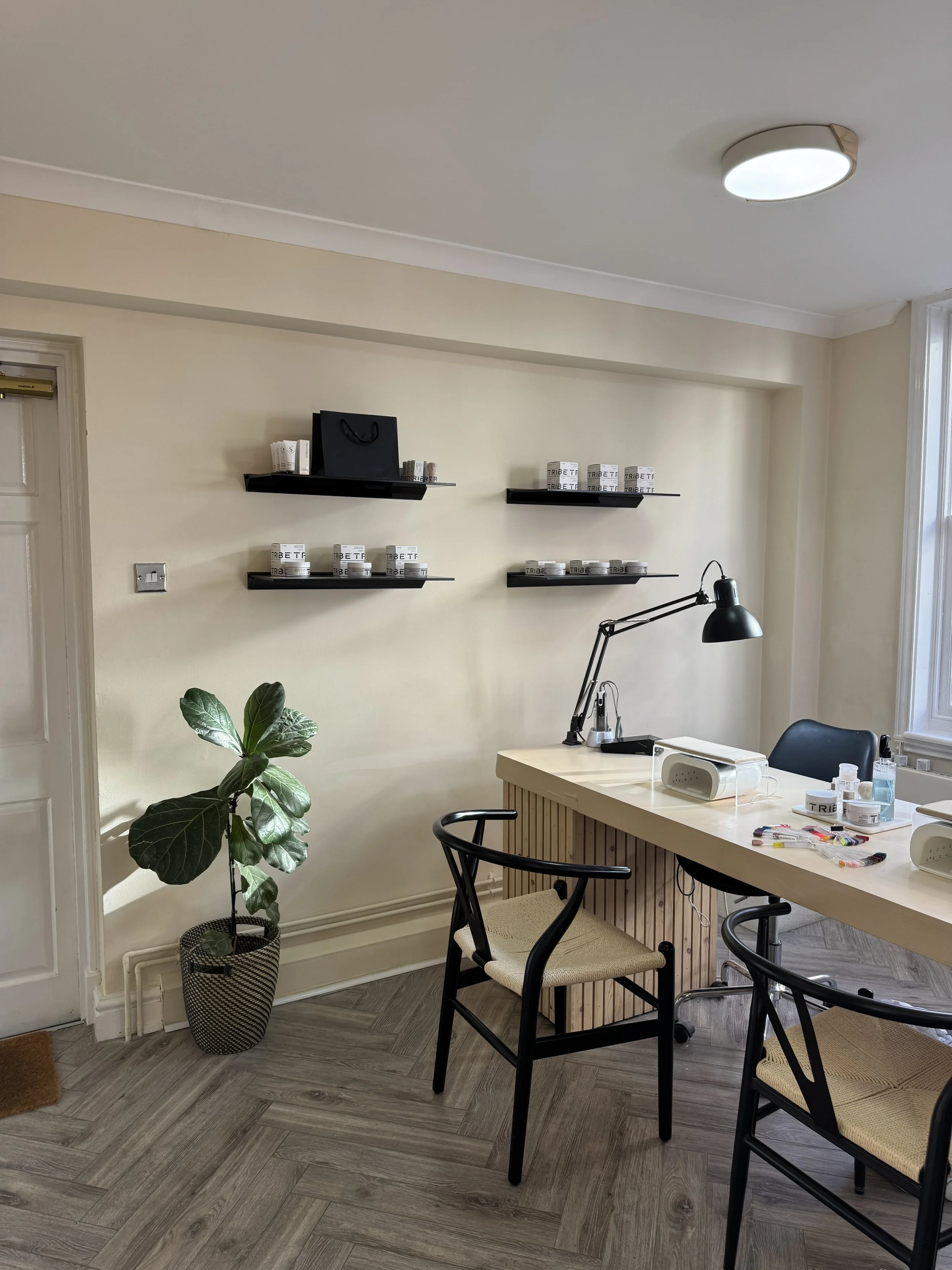 A modern nail salon interior with a clean and minimalist design. The room features a wooden manicure table with two chairs, a desk lamp, and manicure supplies. Shelves on the wall hold various pots or containers. A potted plant is placed on the floor