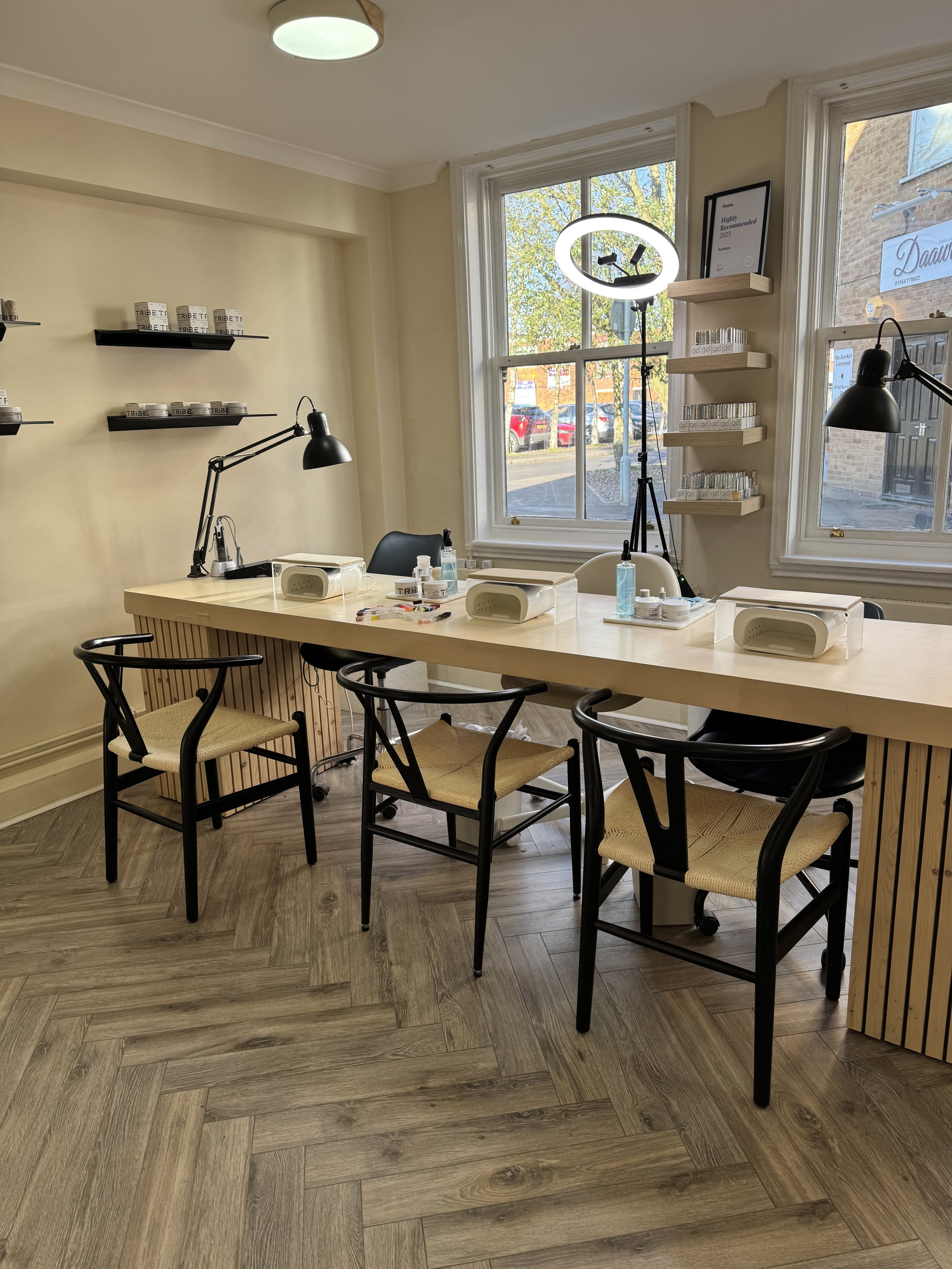 Interior of a nail salon with wooden flooring, a long table, chairs, and manicure stations with lamps, nail supplies, and a ring light near the window.