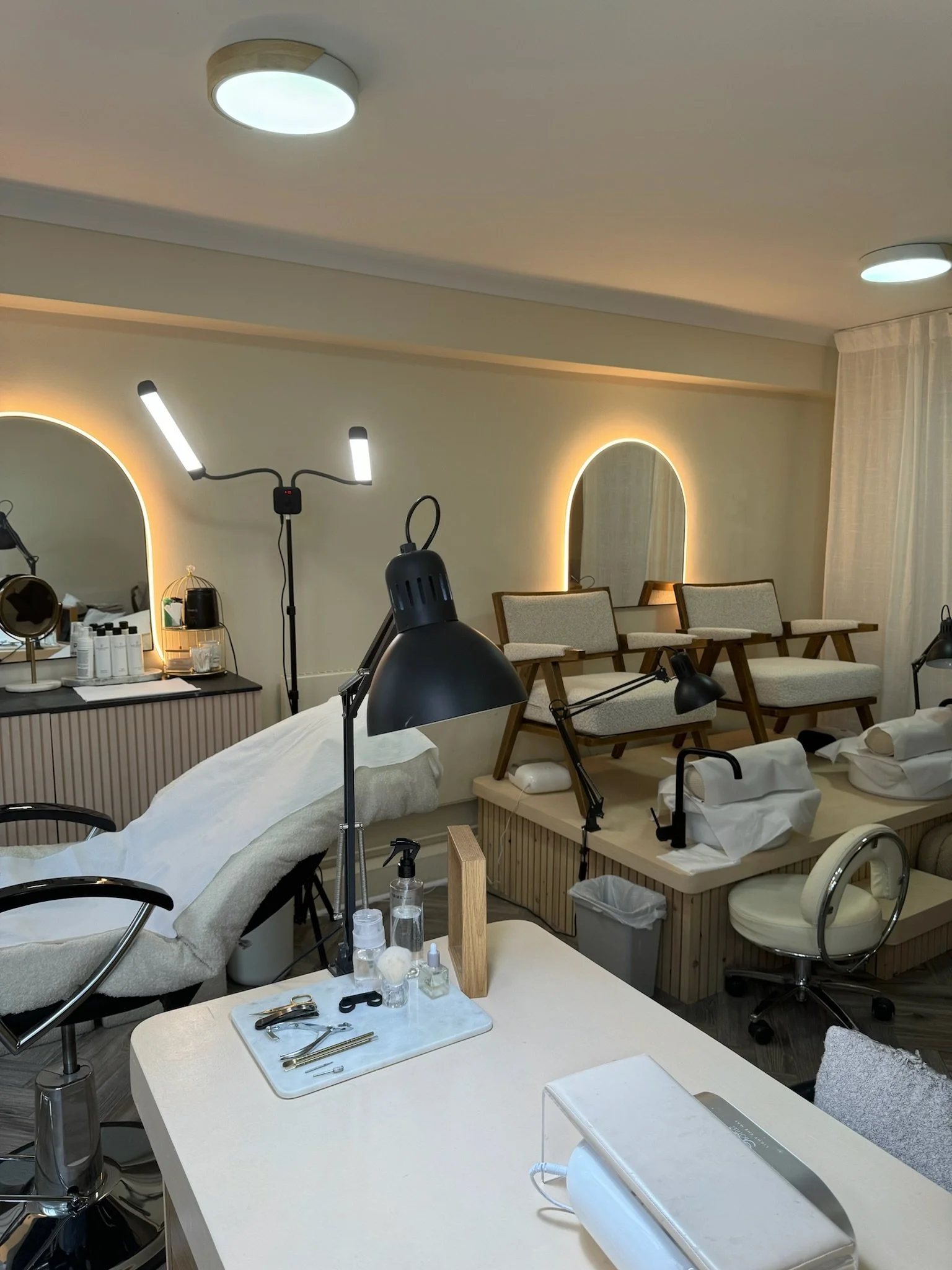 Interior of a modern salon, featuring pedicure stations with cushioned seating and individual lamps, a manicure table with tools and supplies, mirrors with backlighting, and a comfortable treatment chair.
