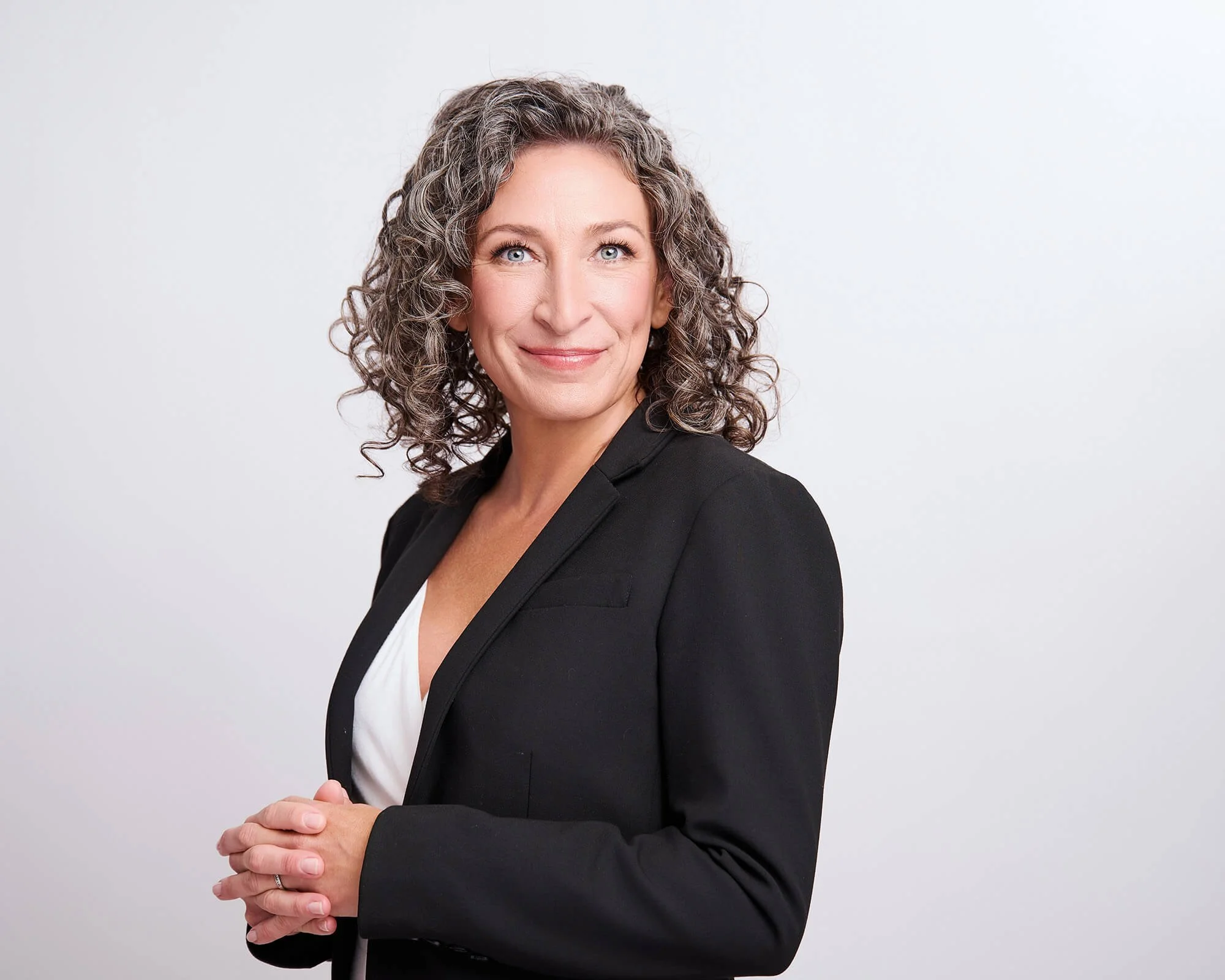 A woman with curly gray hair wearing a black blazer and white top, smiling and looking to the side against a plain white background.