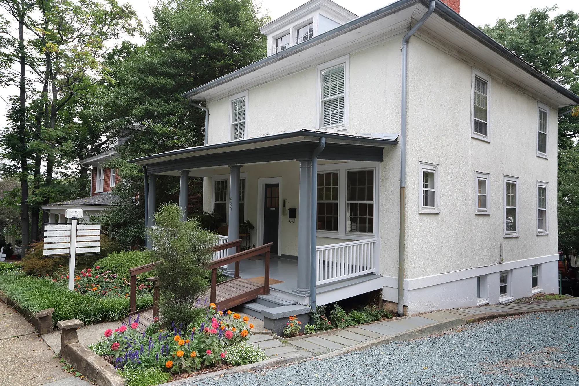White multi-story house with a front porch and flower garden, trees in the background, and a street sign reading 420 in the foreground.