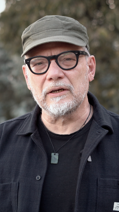 Hombre mayor con barba y gafas negras, usando una gorra y ropa oscura, en un entorno al aire libre.