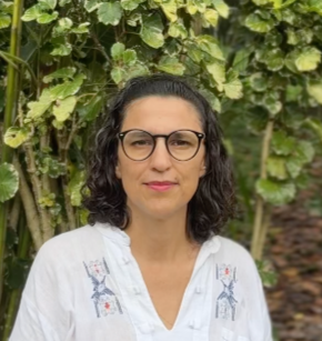 Retrato de una mujer con cabello negro, piel clara, vistiendo una camiseta de rayas blancas y azules, de fondo natural con plantas verdes.