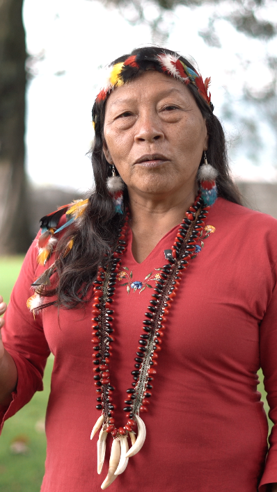 Mujer indígena con atuendo tradicional, collar de huesos y pendientes, en un entorno al aire libre.