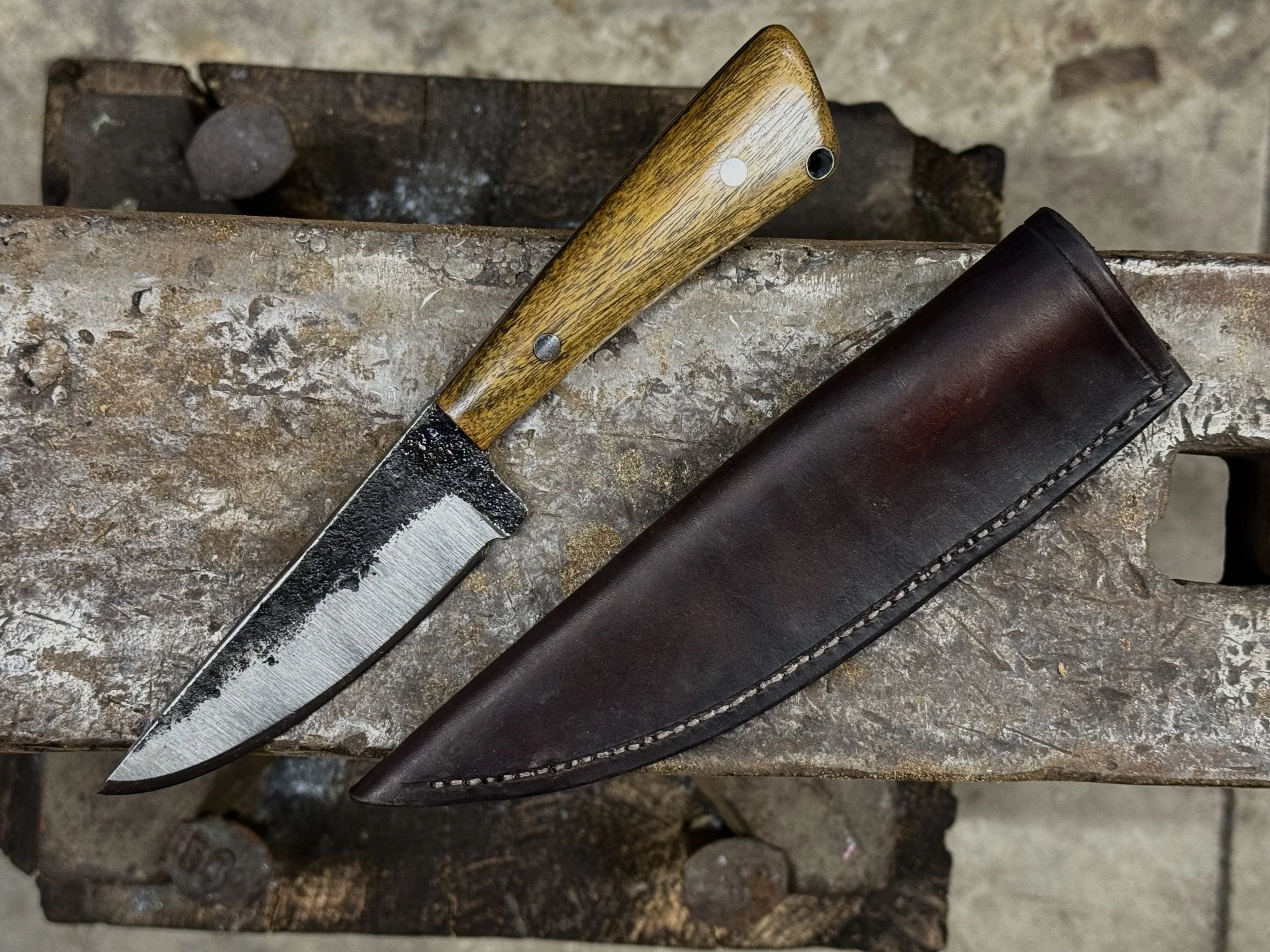 A fixed-blade knife with a wooden handle and black bolster, resting on a rusty metal work surface next to a dark leather sheath.