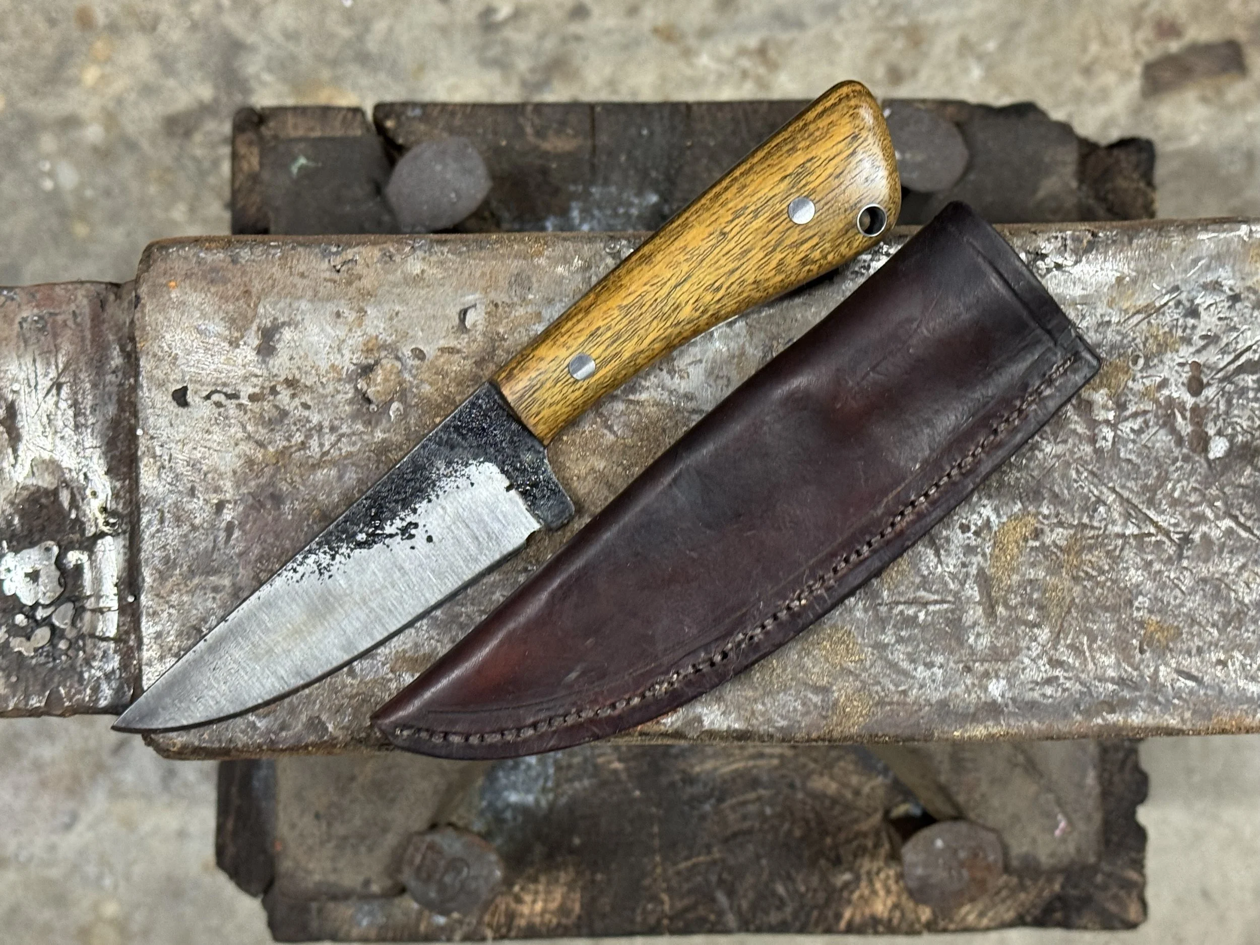 A worn fixed-blade knife with a wooden handle and a leather sheath on a rusty workbench.