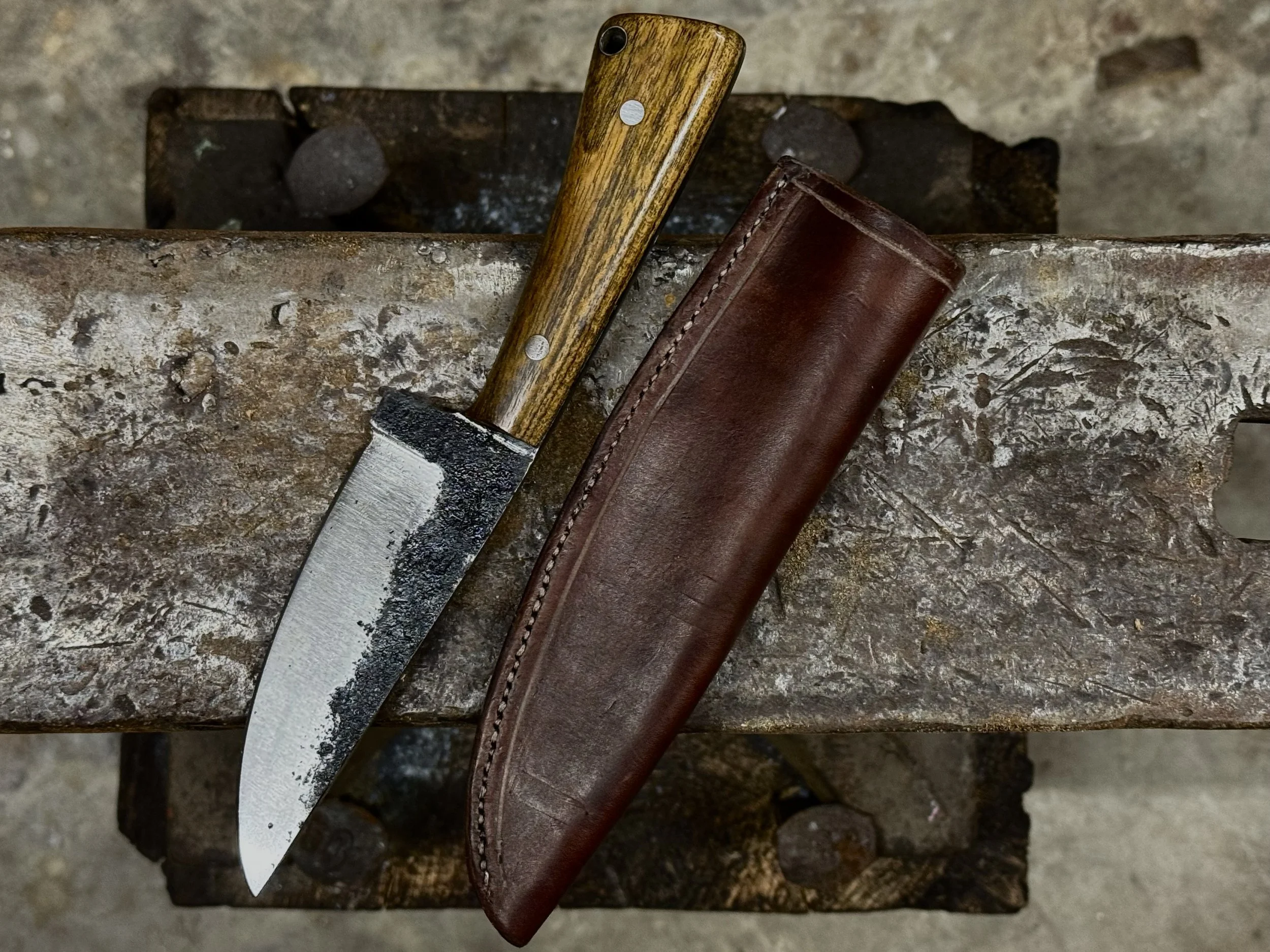 A hunting or utility knife with a wooden handle, a black and silver blade, and a brown leather sheath, resting on a rusty anvil.