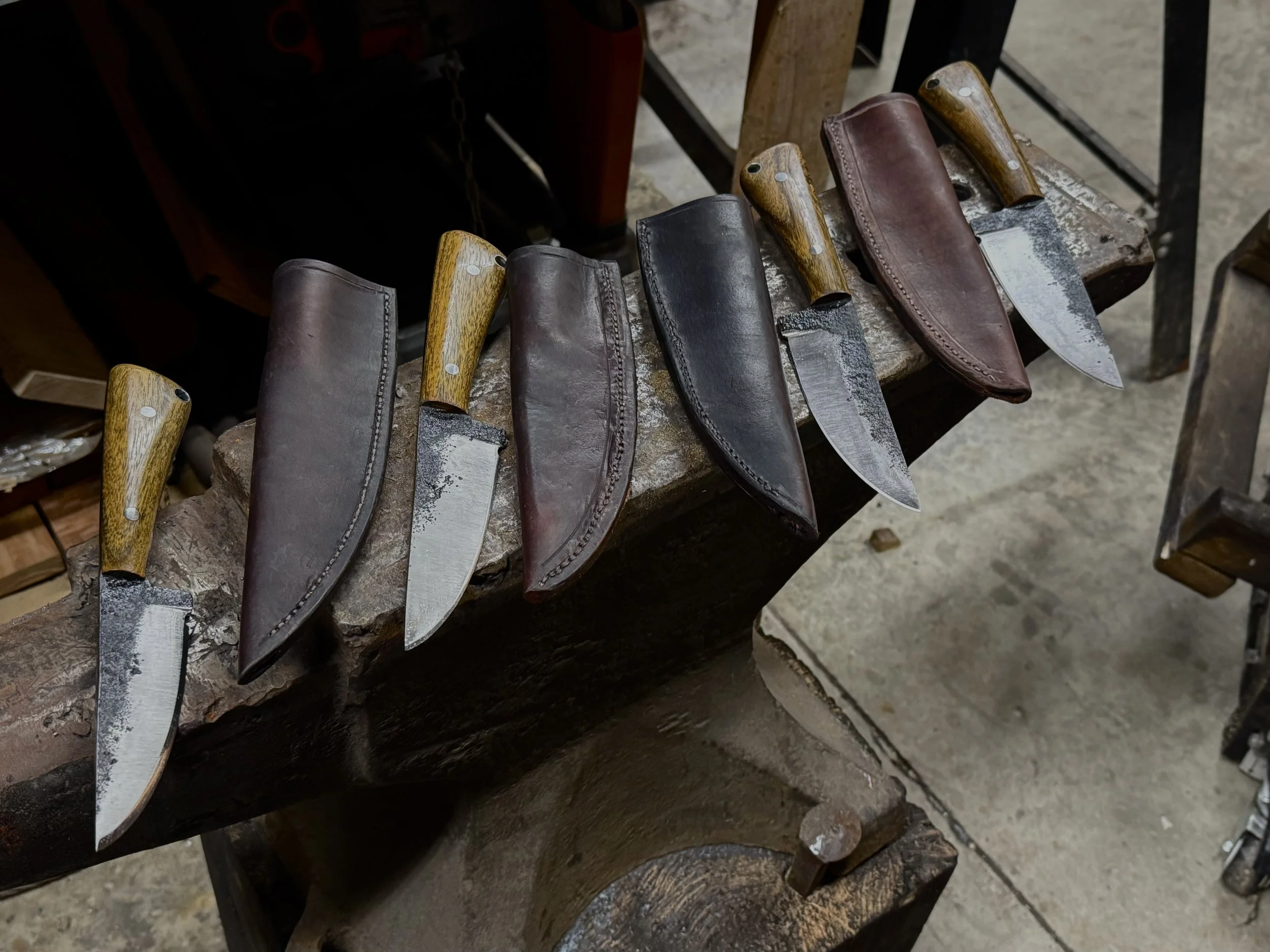 Set of handcrafted knives with wooden handles, leather sheaths, and metal blades, displayed on a workbench in a blacksmith workshop.