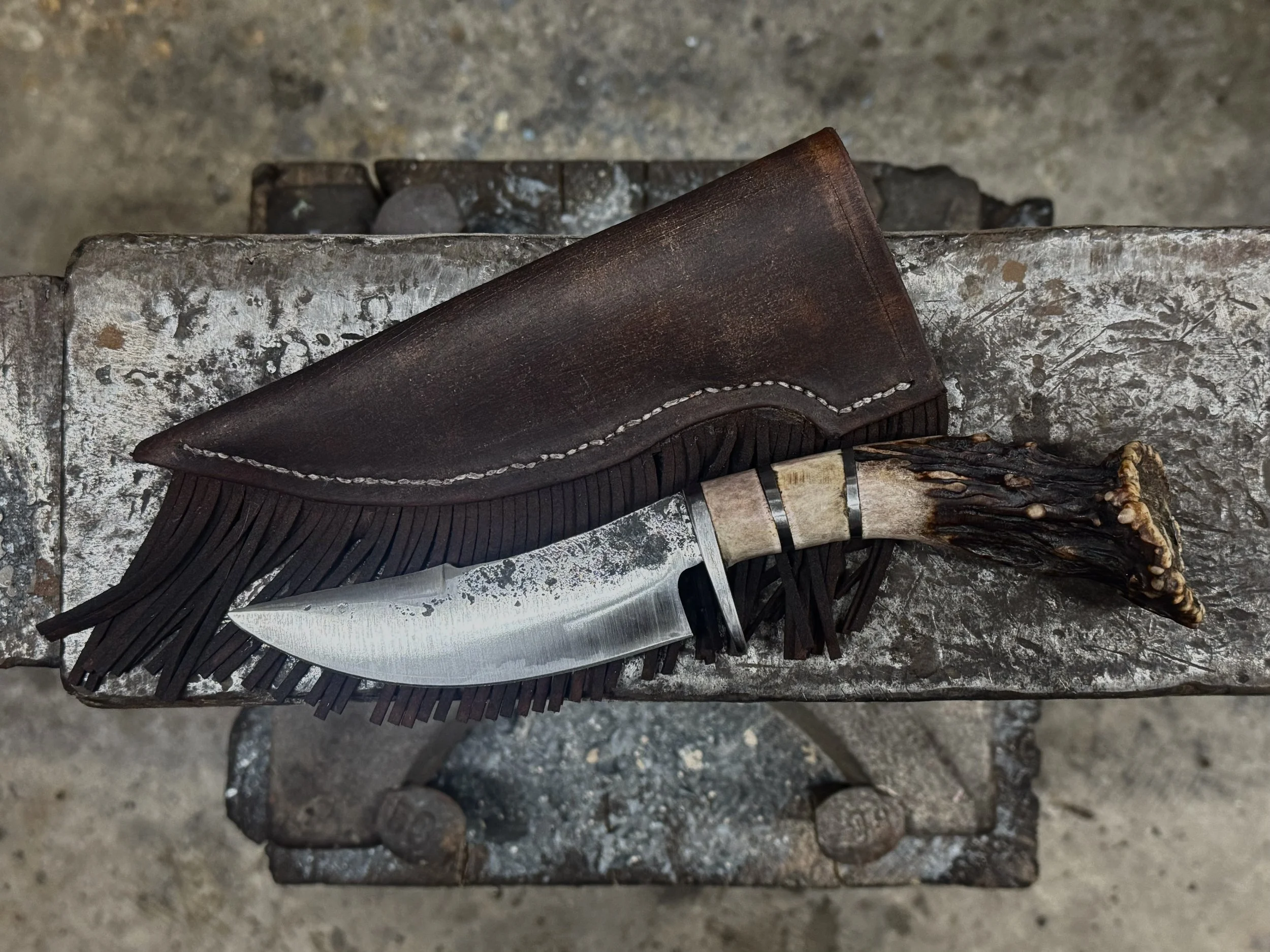 A hunting knife with a curved stainless steel blade and an antler handle, placed on a leather sheath with fringe, on a worn metal surface.