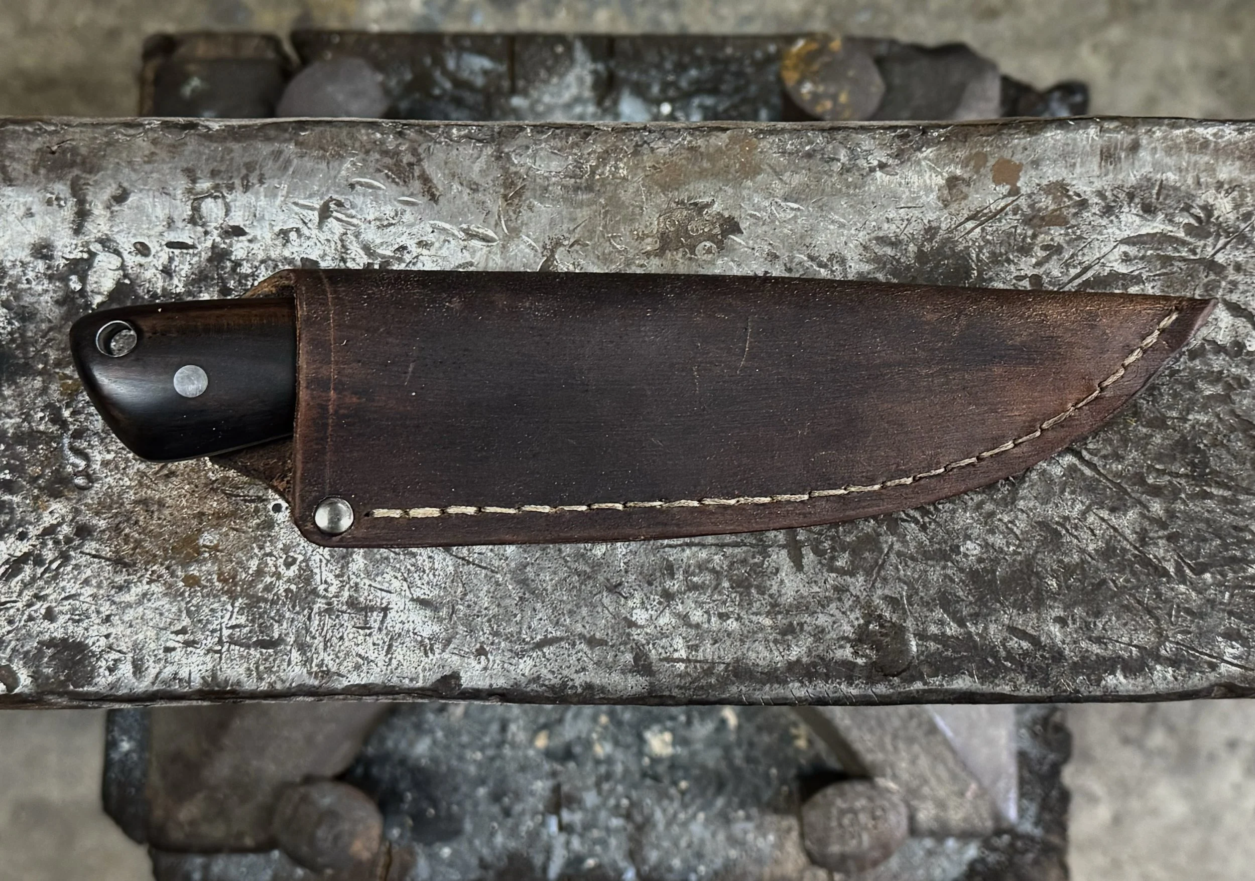 A black-handled knife with a dark brown leather sheath, lying on a rough, weathered metallic surface, with a blurry metallic structure underneath.
