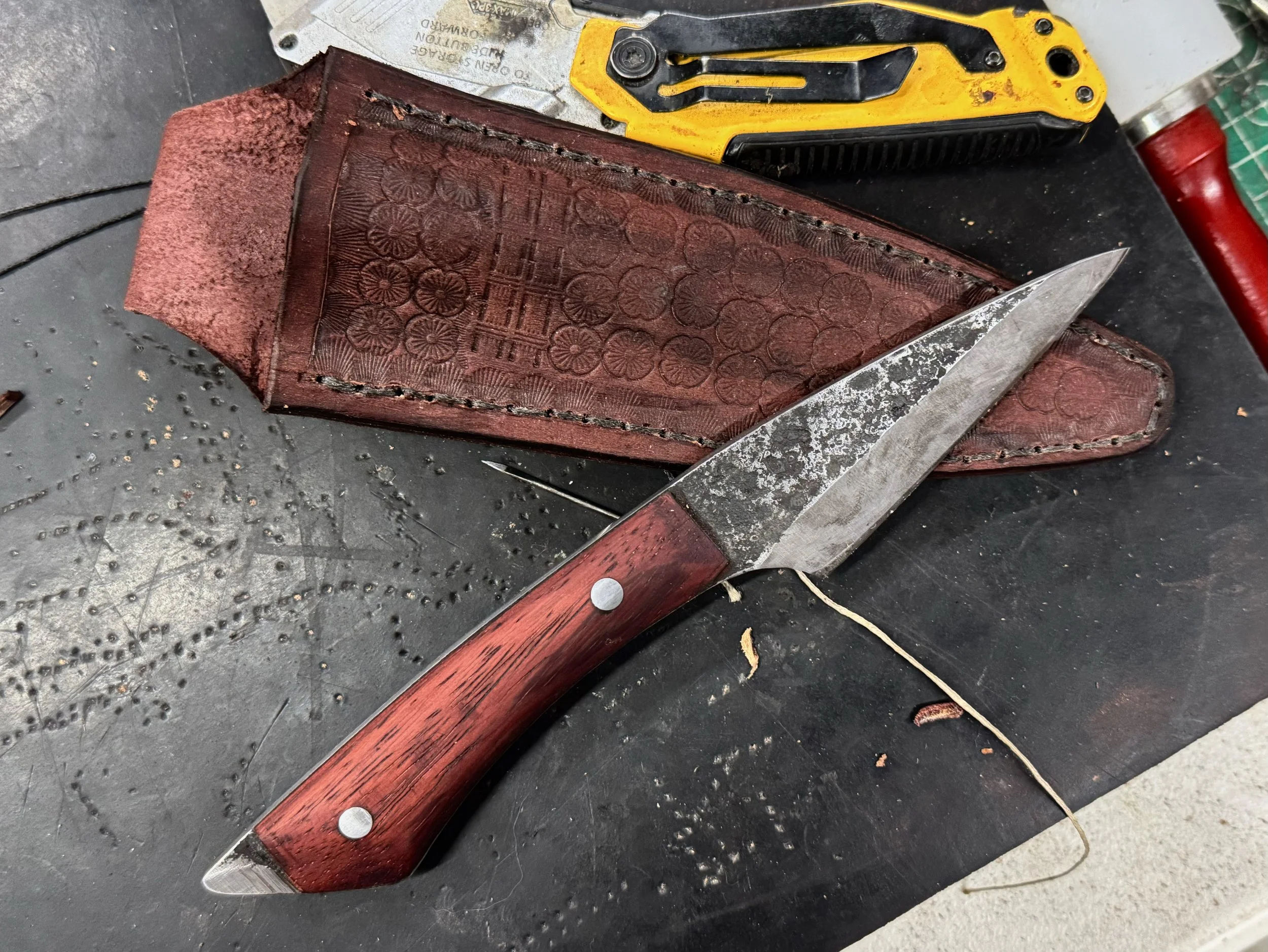 A worn hunting knife with a red wooden handle and a partially rusted blade, resting on a black surface along with a brown leather sheath and various tools.