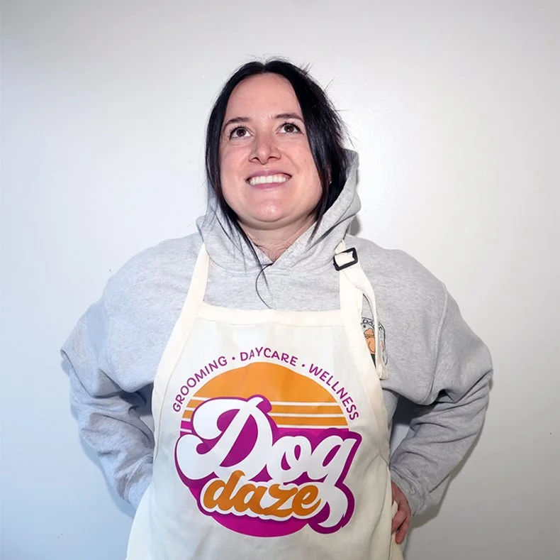 A woman smiling and wearing a gray hoodie and a white apron with the logo "Dog Daze" which offers grooming, daycare, and wellness services for dogs, standing against a plain white wall.