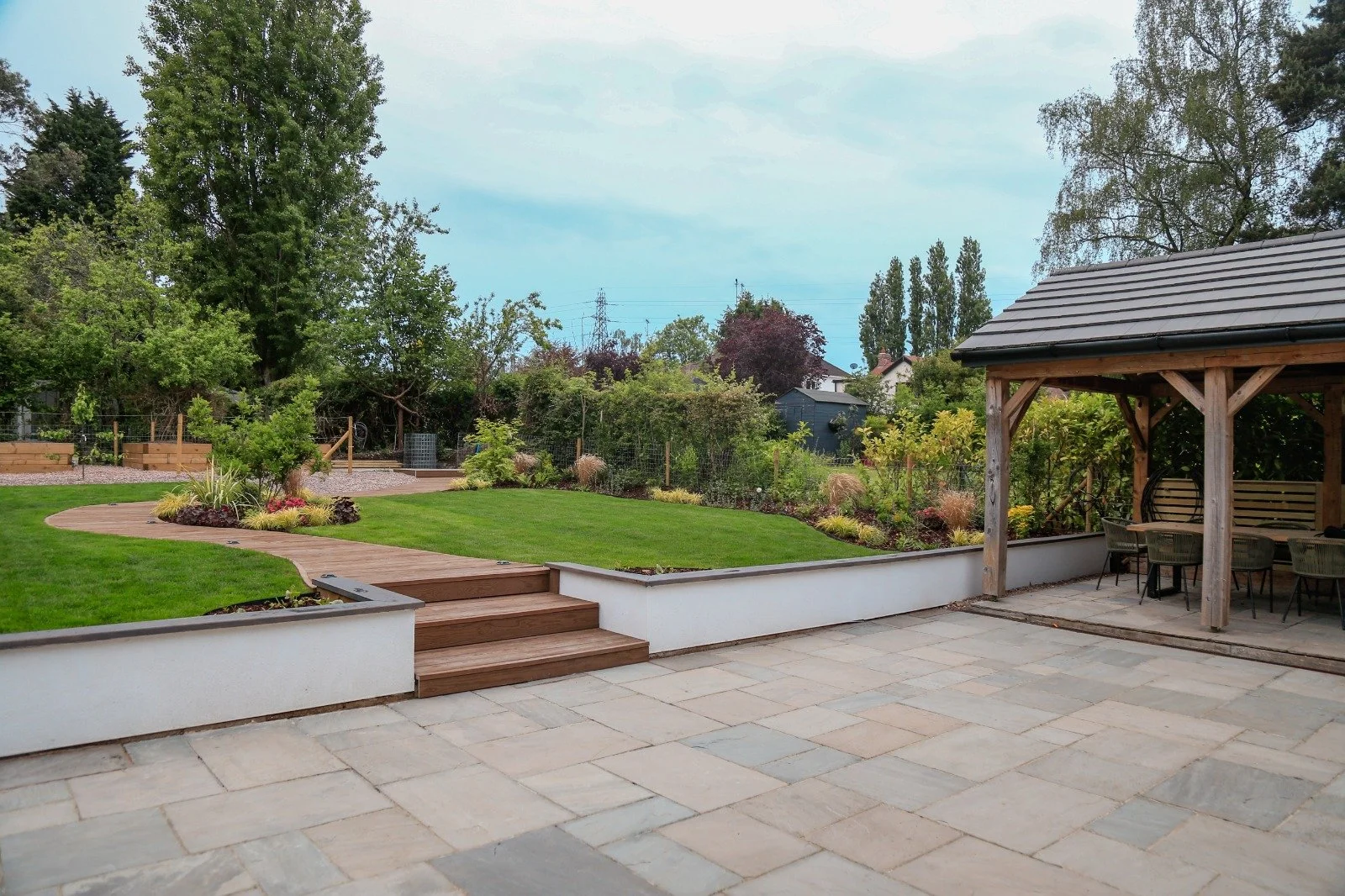 A landscaped garden in Warwickshire, with a stone patio, wooden steps leading to a grassy area, and a covered seating area with tables and chairs. There are flower beds and shrubbery along the perimeter.