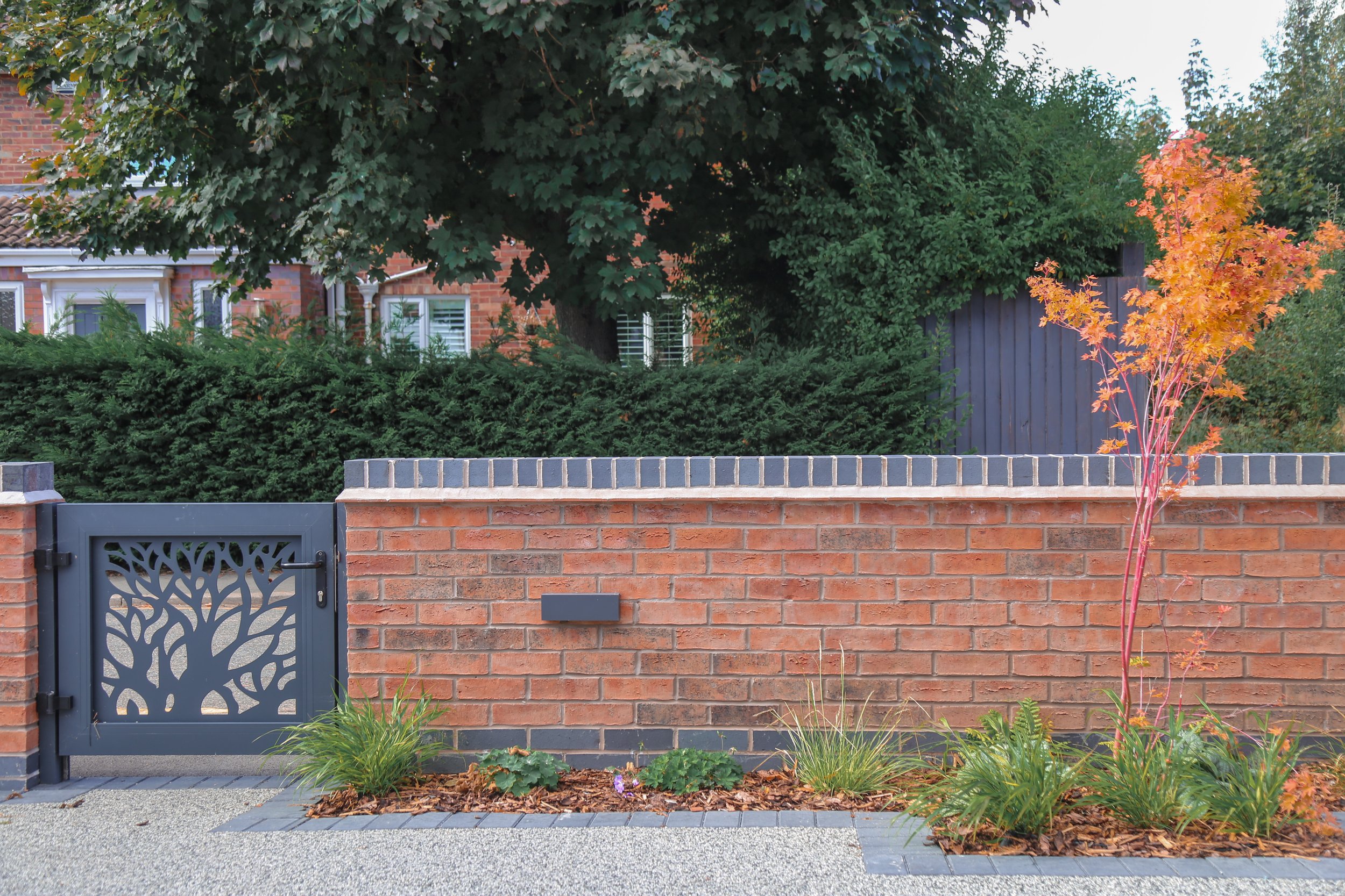 Brick wall with a gate featuring a tree design, small plants and mulch in front, green bushes, a large tree, a dark-colored fence, and a house with white-framed windows in the background.