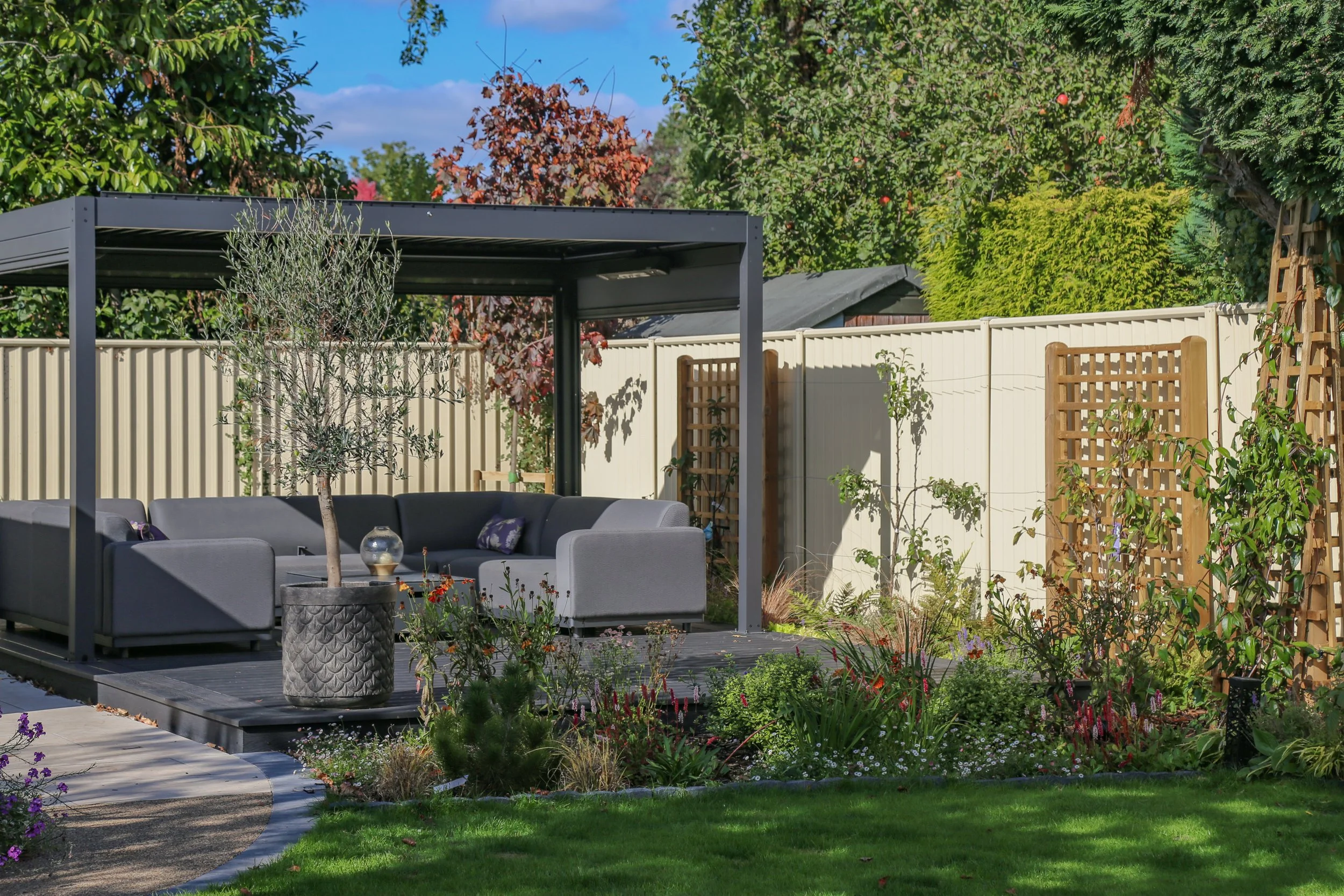 A Well designed outdoor area, with an aluminium pergola, sitting on a composite deck , surrounded by planting beds
