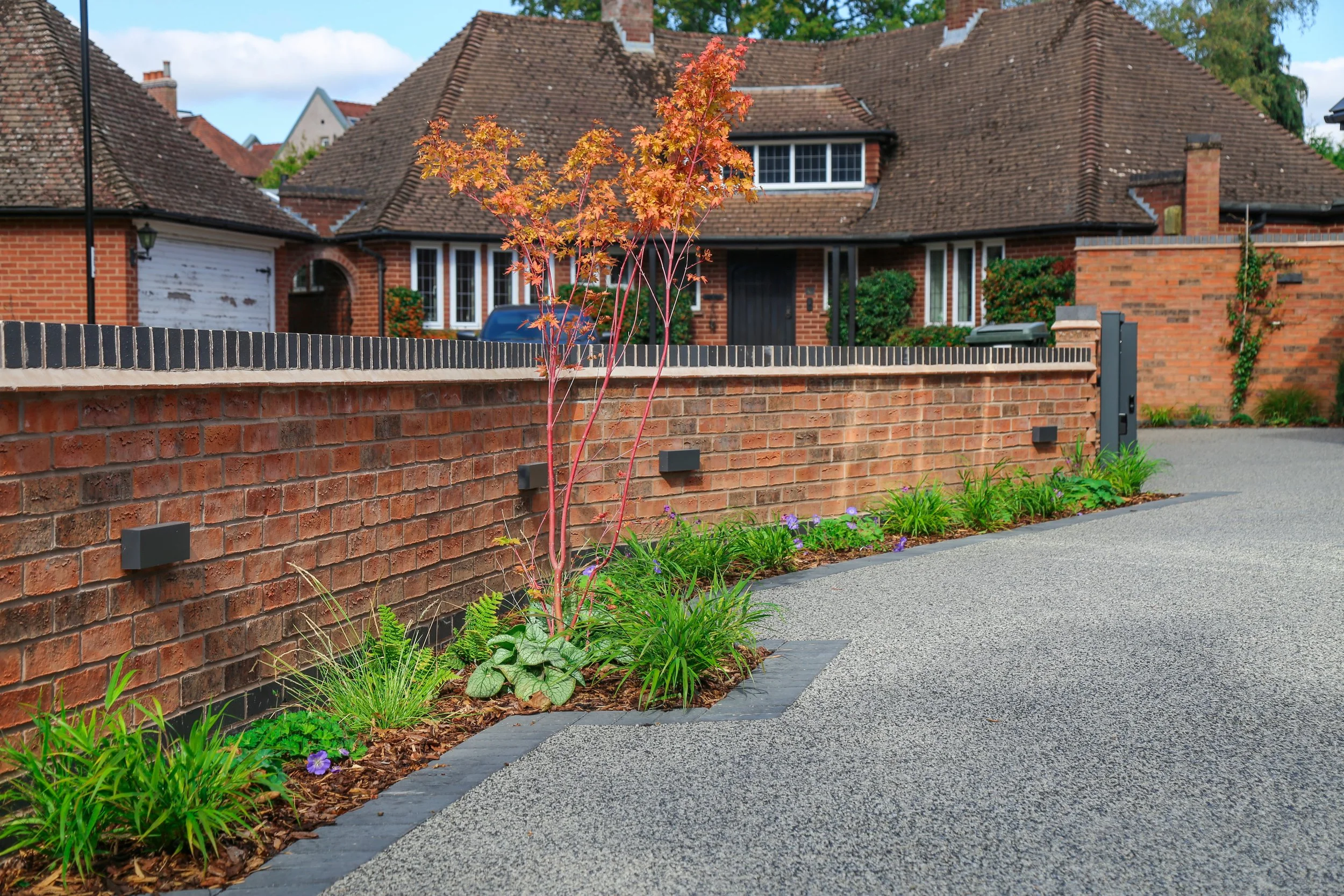 A residential neighborhood with a brick wall, landscaped garden bed, and a house in the background. The garden includes a small red-leafed tree and green plants with purple flowers. The street is clean with gray pavement.