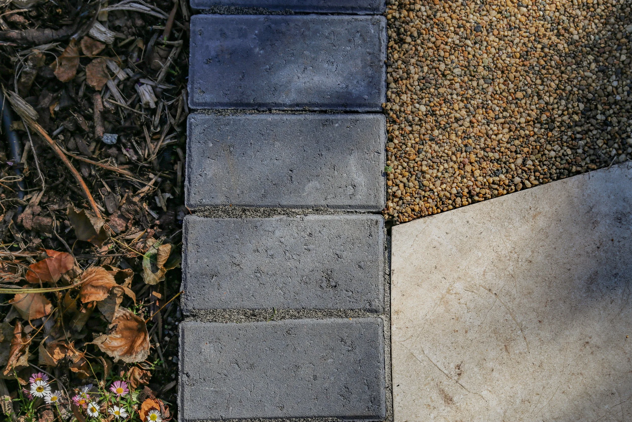Close-up of a sidewalk showing concrete pavers, gravel, and a section of dry leaves and small flowers.