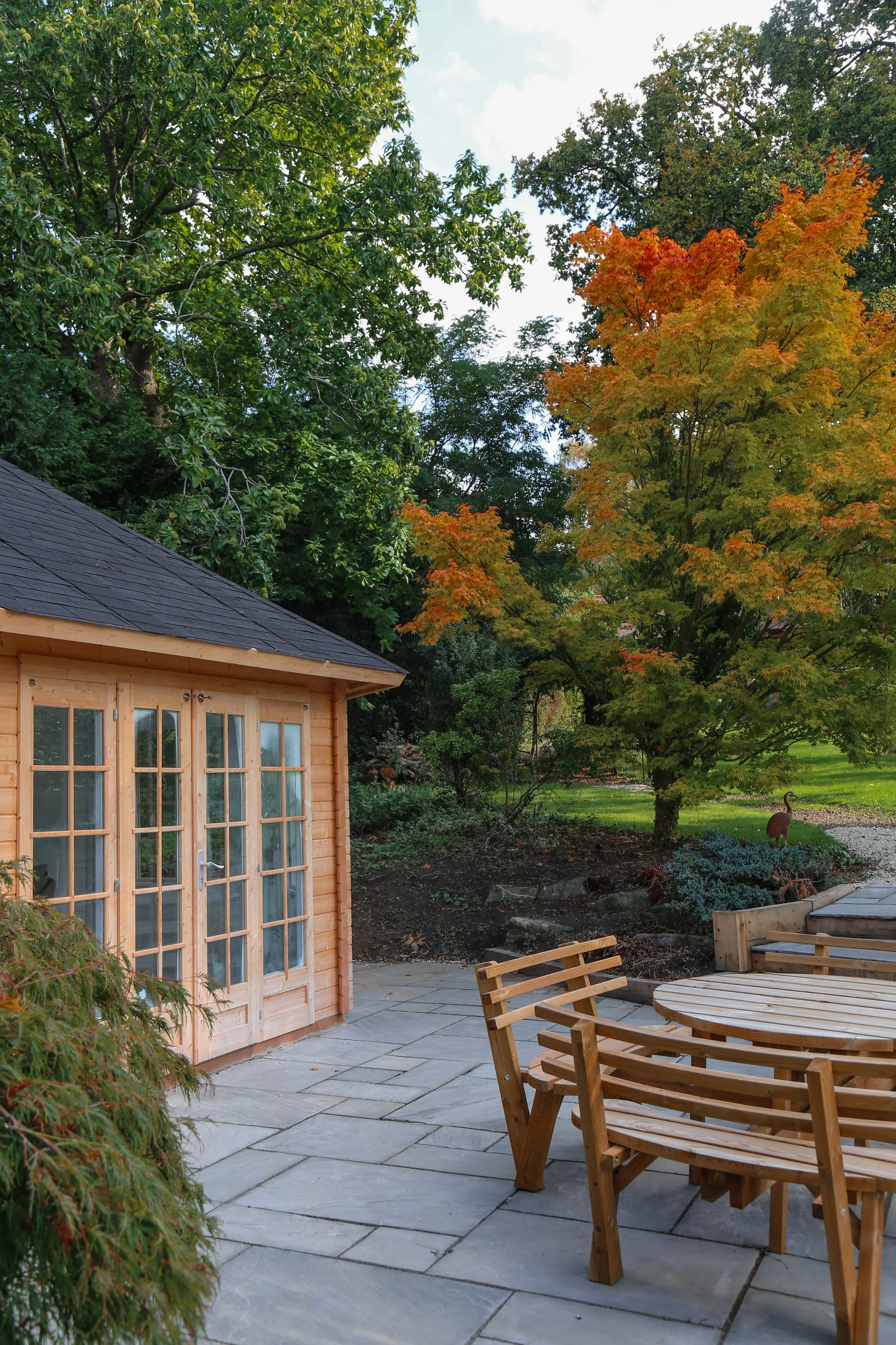 natural stone patio, with a large wooden outdoor garden room and a table and chairs, autumn leaves on the trees surrounding