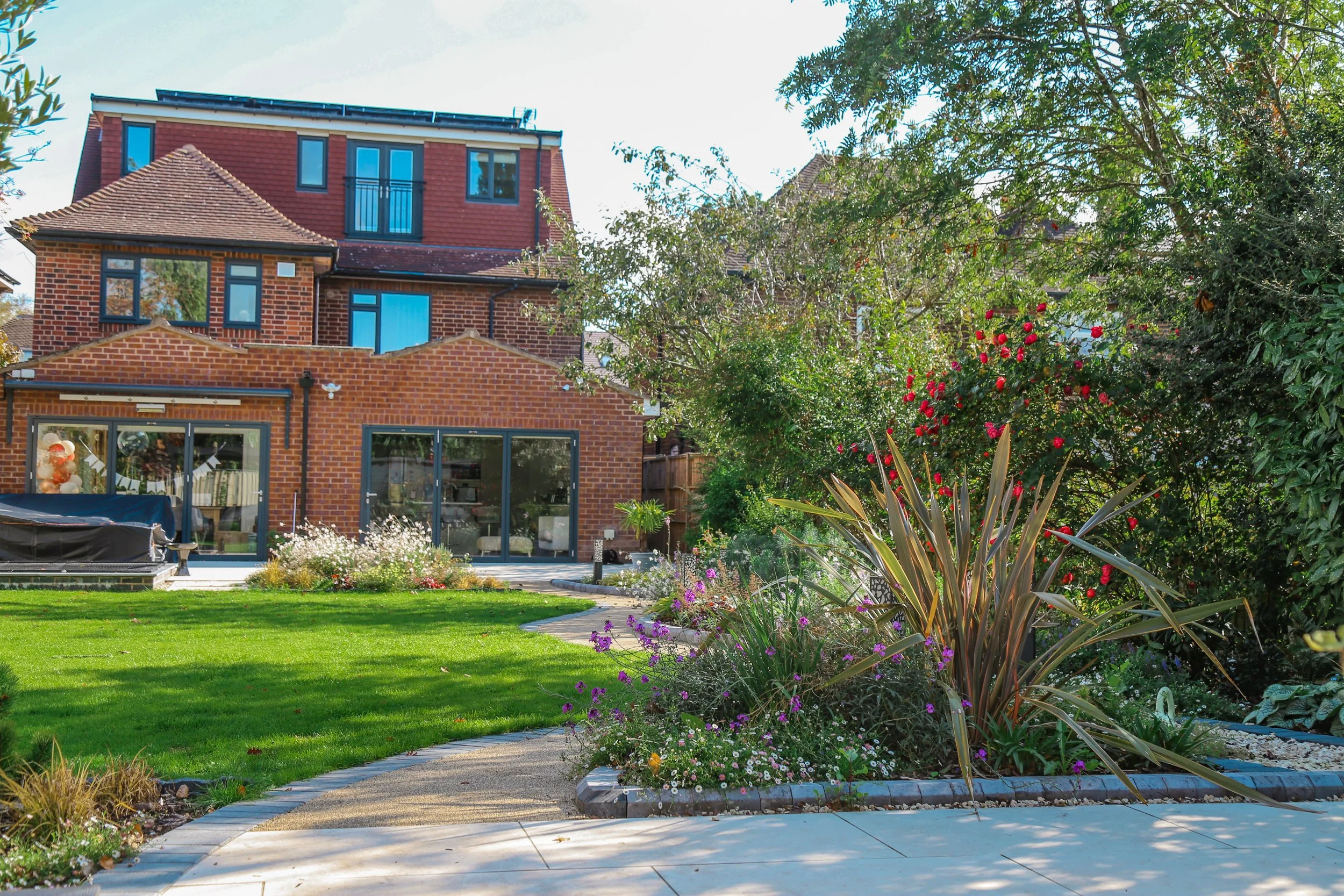 A back garden with a manicured grassy lawn, colorful flowers, bushes, and trees, with a brick house in the background.