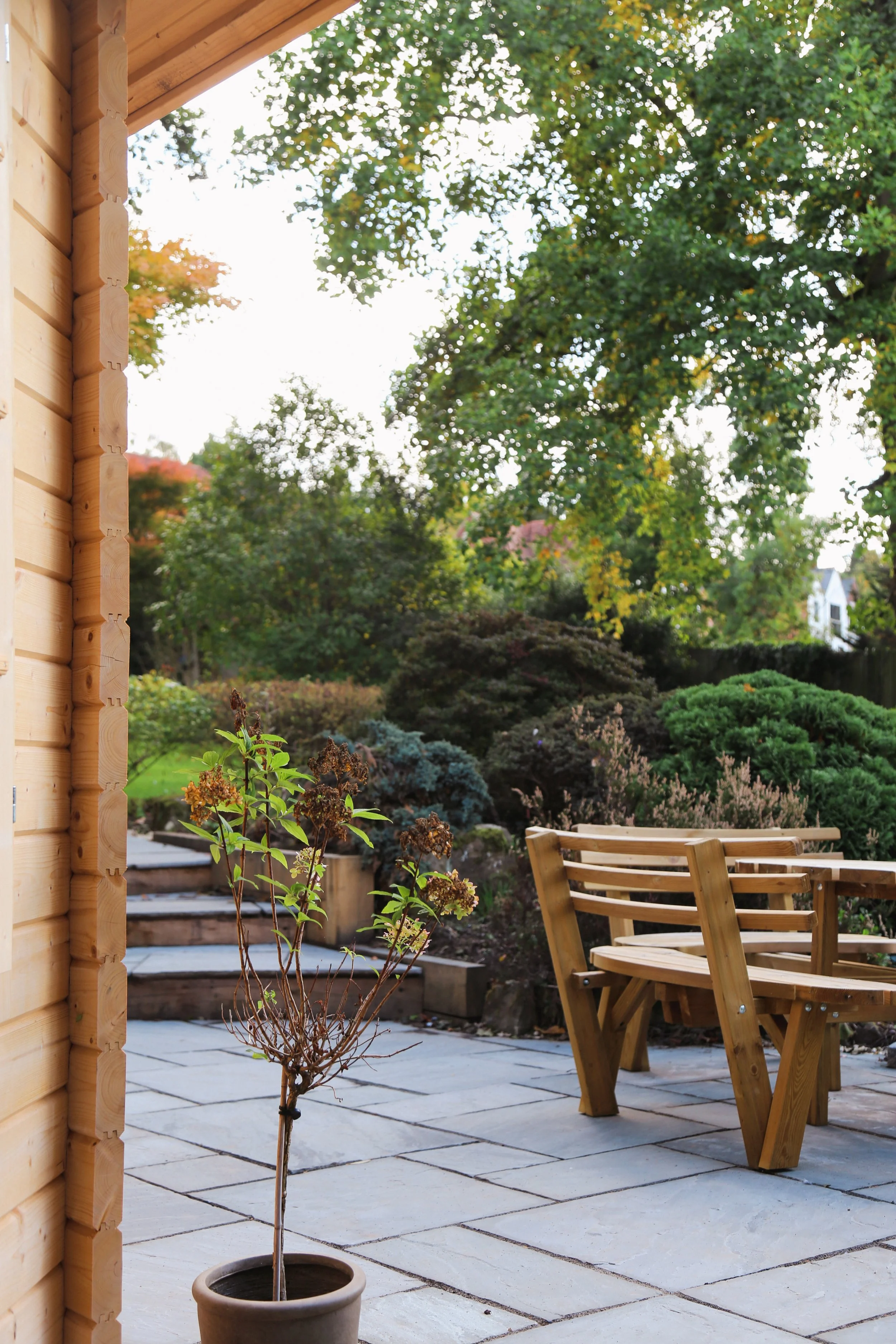 A natural stone patio next to a wooden garden  building and mature planting beds