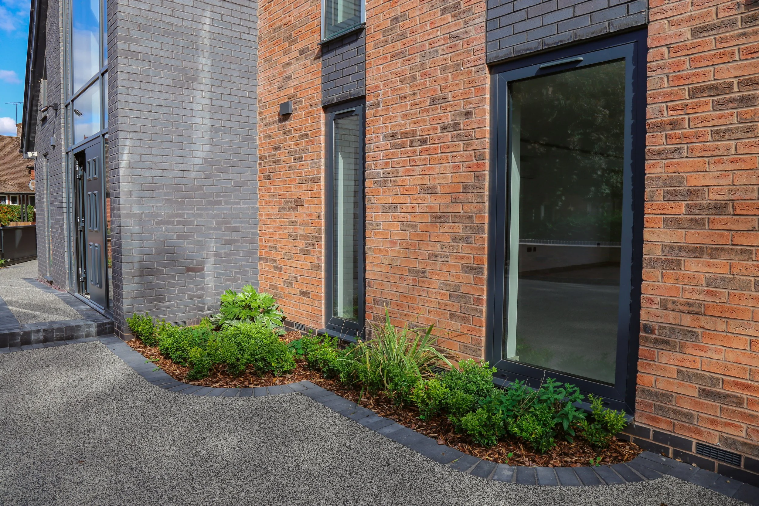 Modern building exterior with gray and red brick walls, large windows, and a landscaped flower bed with green plants and mulch.