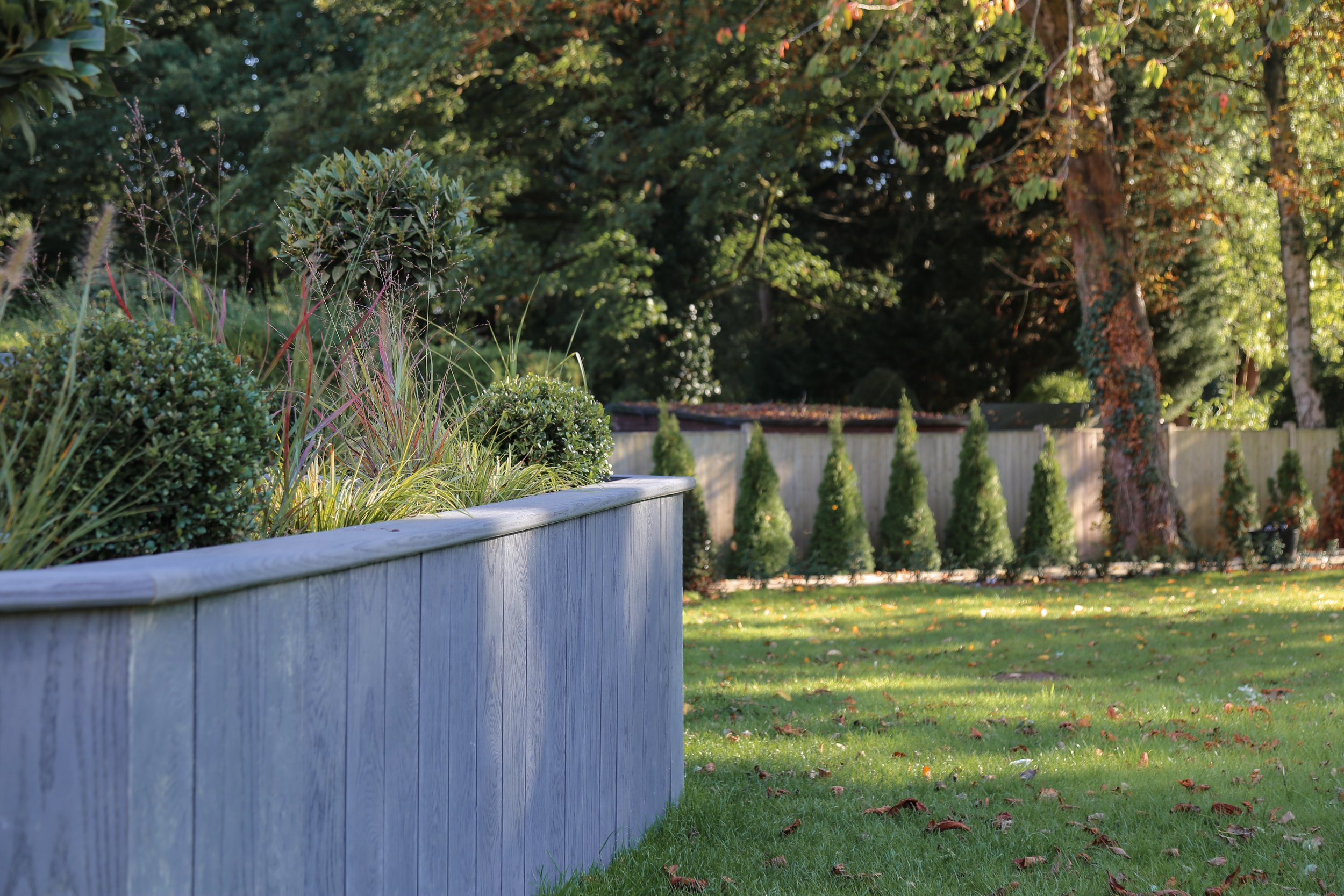 Large raised planting bed with ornamental trees and bushes planted, surrounded by a planted lawn and trees in the background