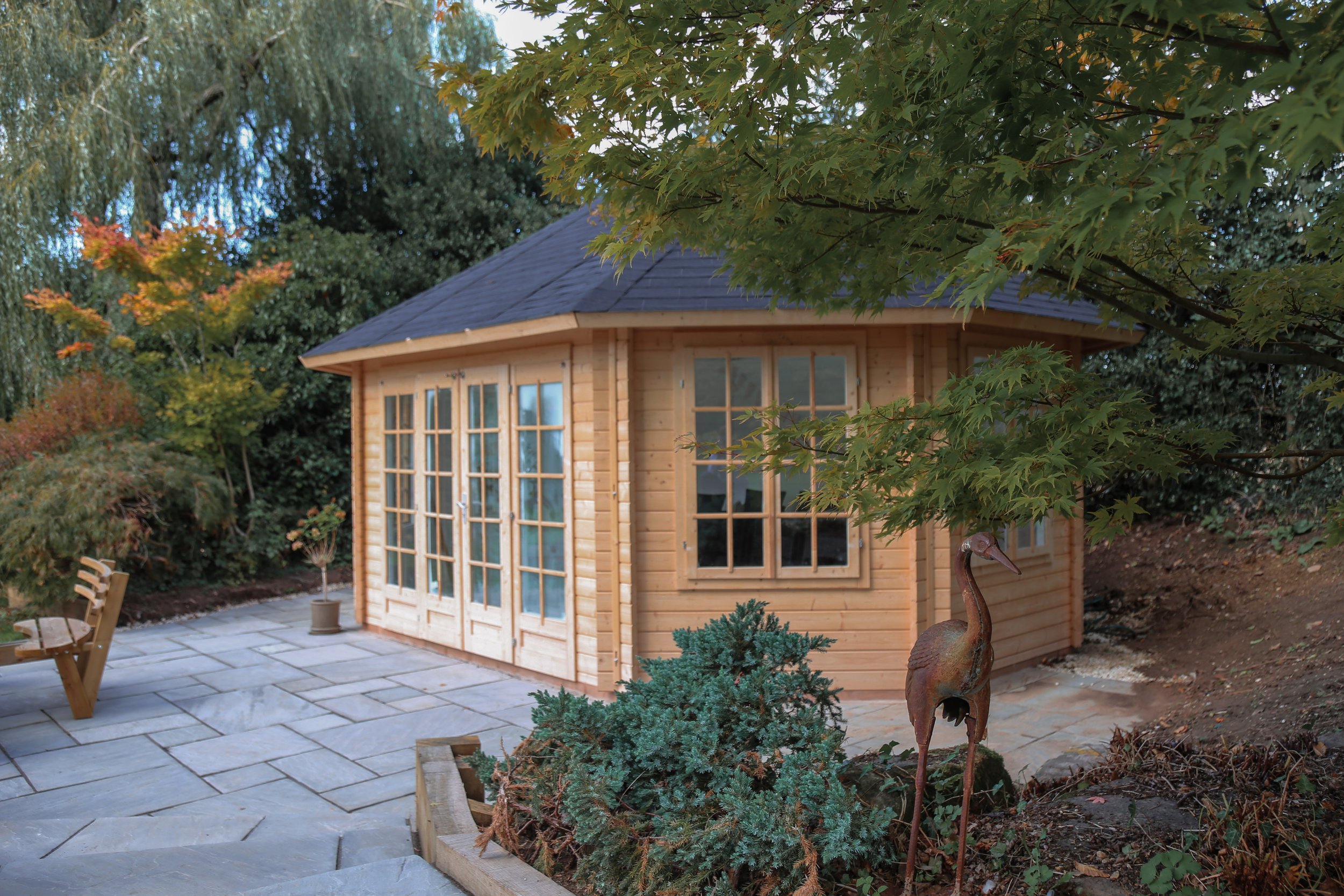 A large garden room surrounded by natural stone paving and lush green planting, a corten steel bird structure sits amongst the planting