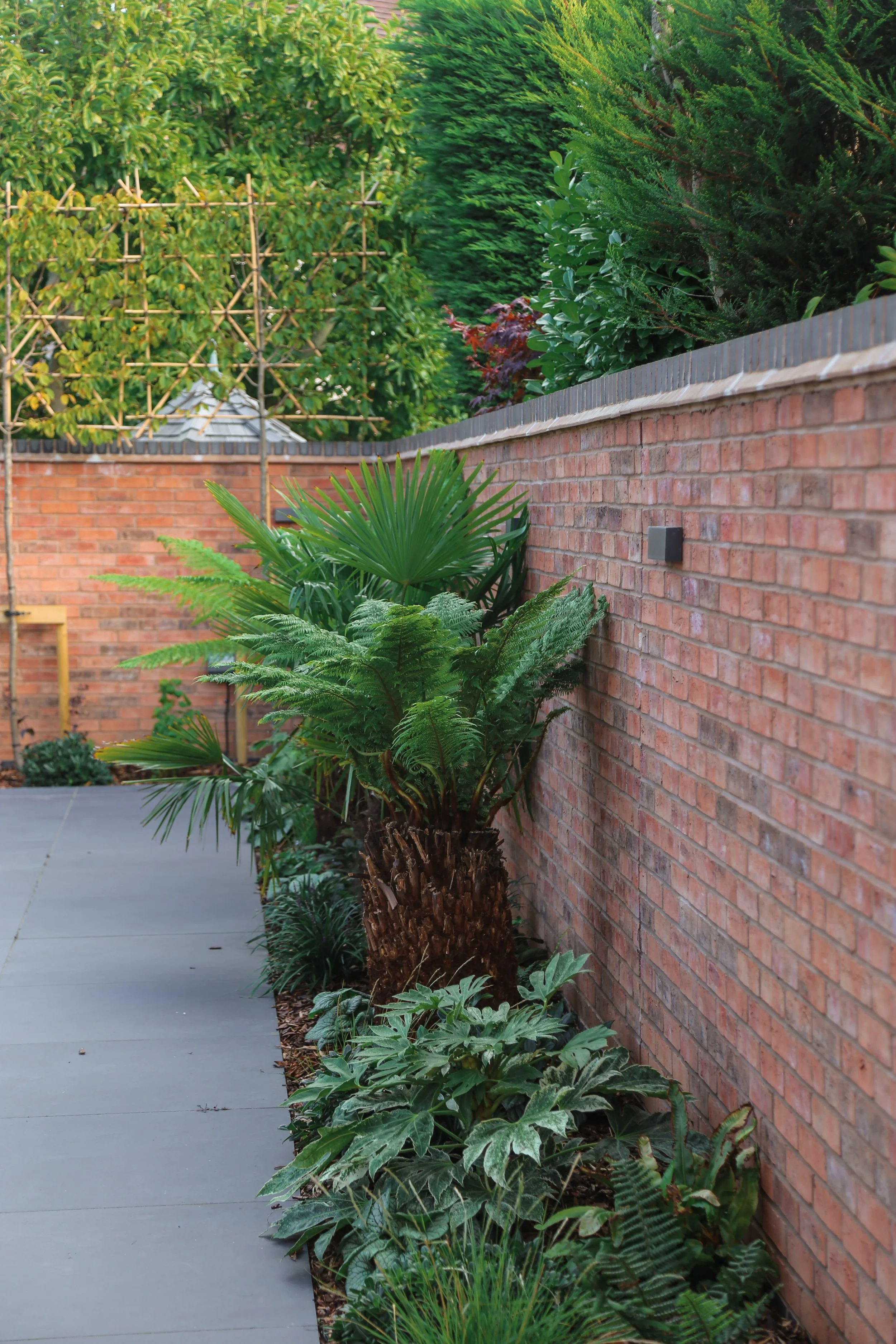 A garden with a brick wall, green plants, and trees, including ferns, palms, and shrubs. Landscaping In Warwickshire