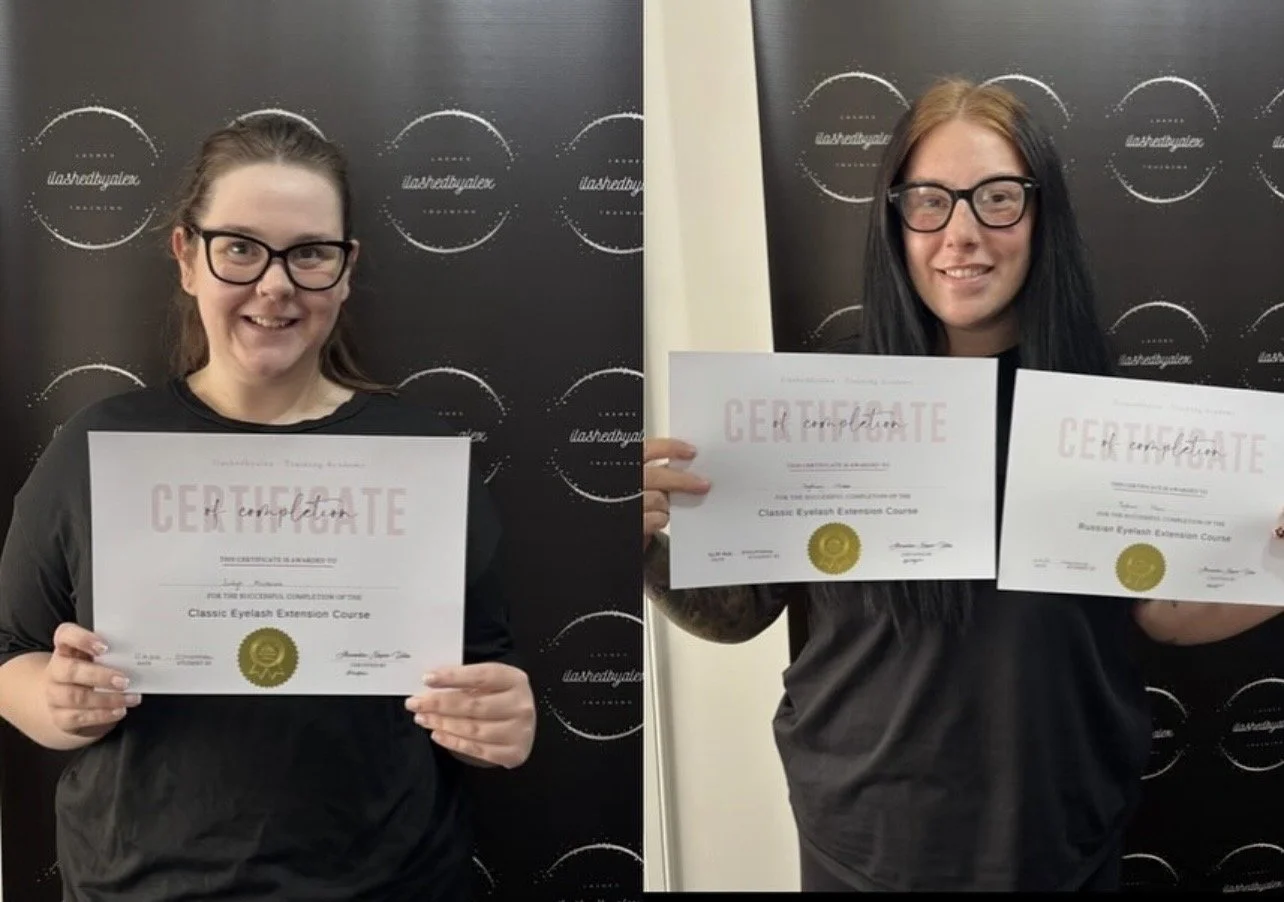 Two women each holding a certificate, standing in front of a black backdrop with white text and logo. The woman on the left has brown hair, glasses, and is wearing a black shirt. The woman on the right has long dark hair, glasses, and is also wearing a black shirt. Both women are smiling and displaying their certificates. The certificates are for completing a classic eyelash extension course.
