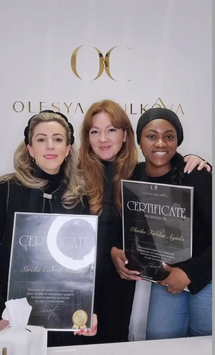 Three women holding certificates in front of a white background with a gold logo and text. They are smiling, wearing black clothing, and appear to be at a celebratory event.