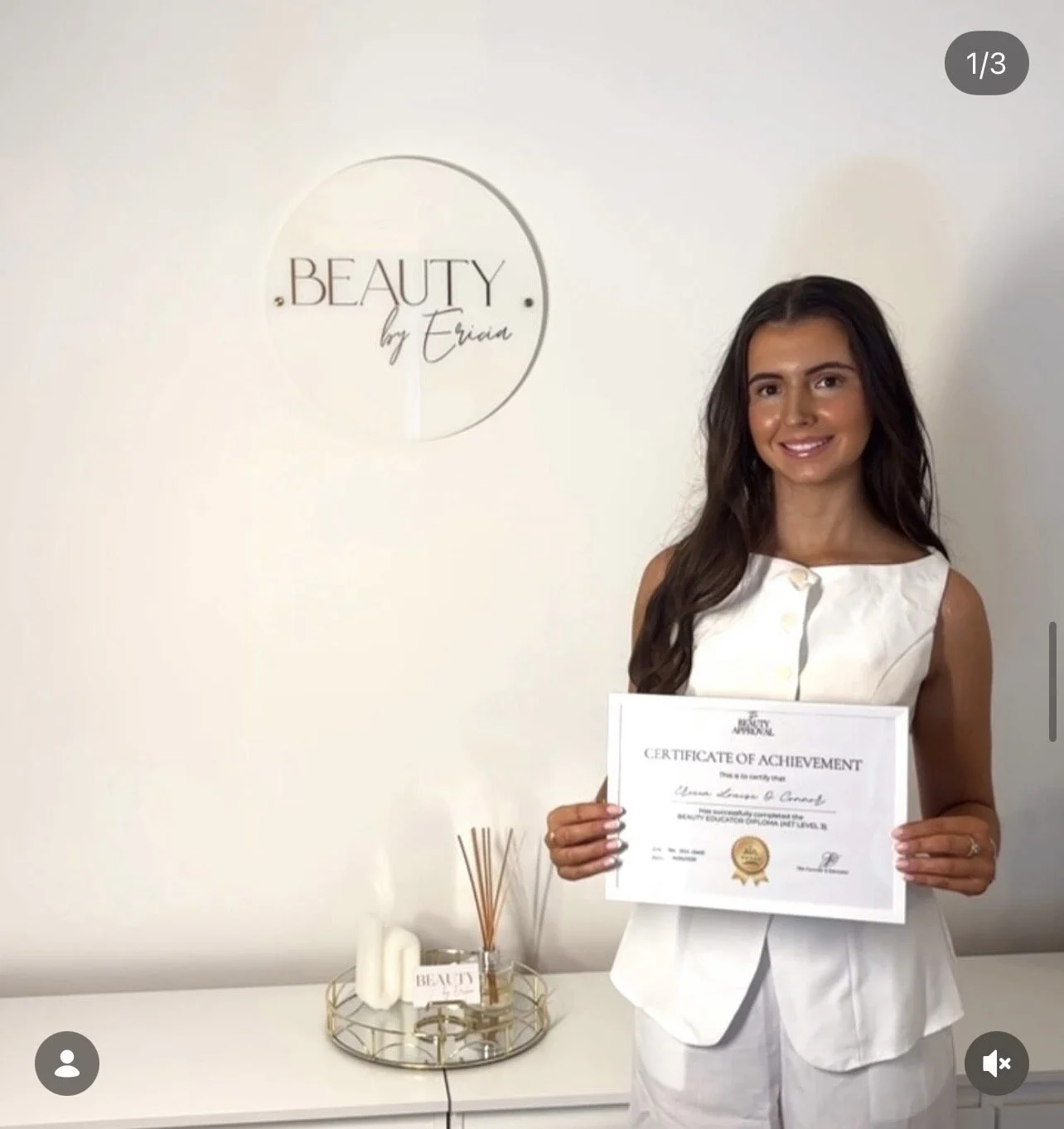 Woman holding a certificate of achievement in front of a white wall with a sign that reads 'BEAUTY by Eria'. A tray with candles and reed diffuser is on the table beside her.