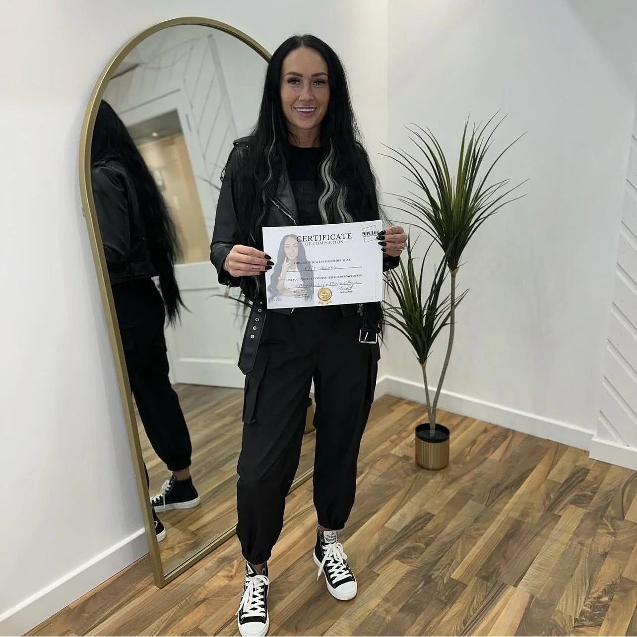 Woman with long dark hair holding a certificate of completion, standing next to a tall potted plant in a room with a wooden floor and a large mirror.