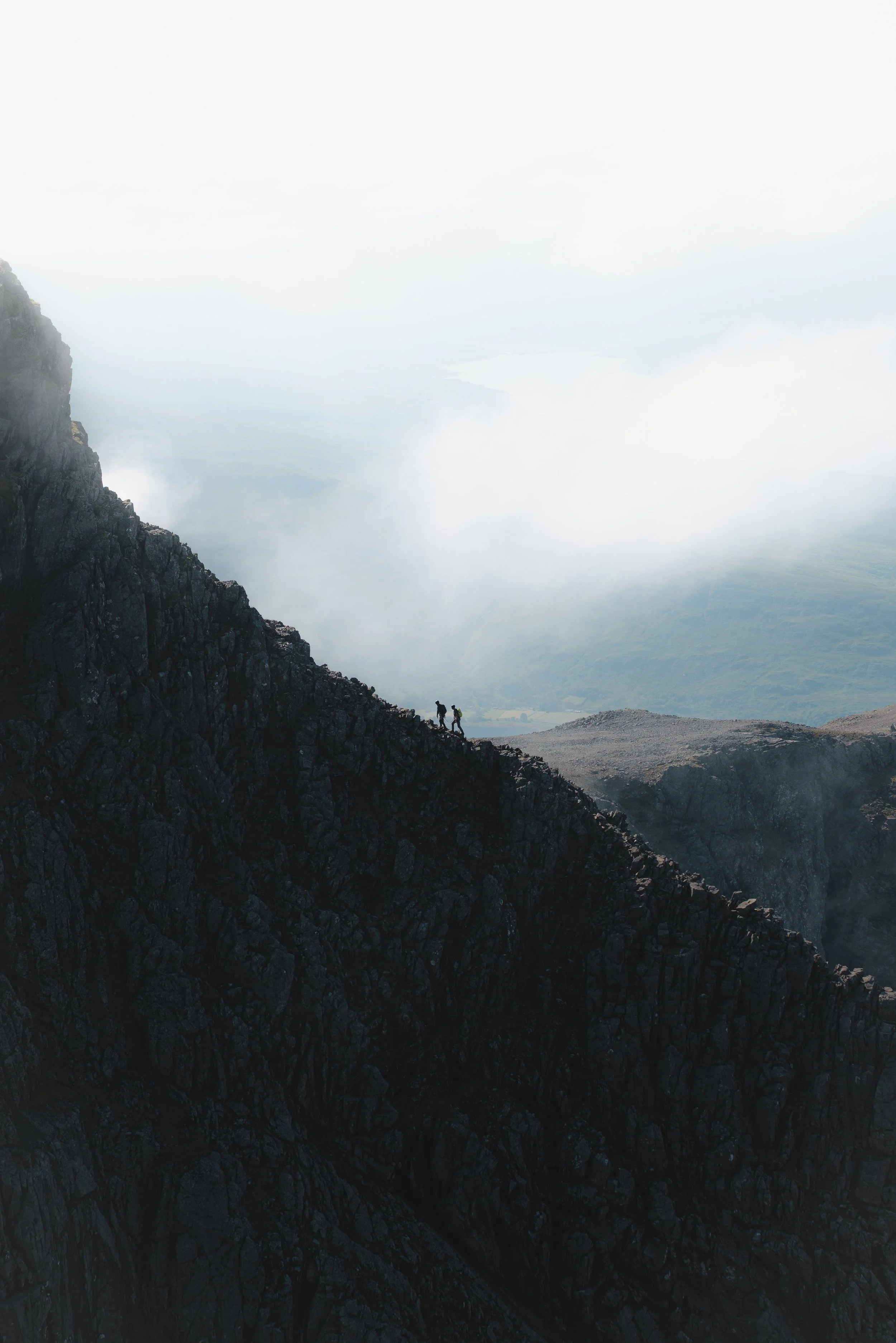 Two hikers walking along a rocky trail on a mountain slope with a foggy, cloud-covered valley below.