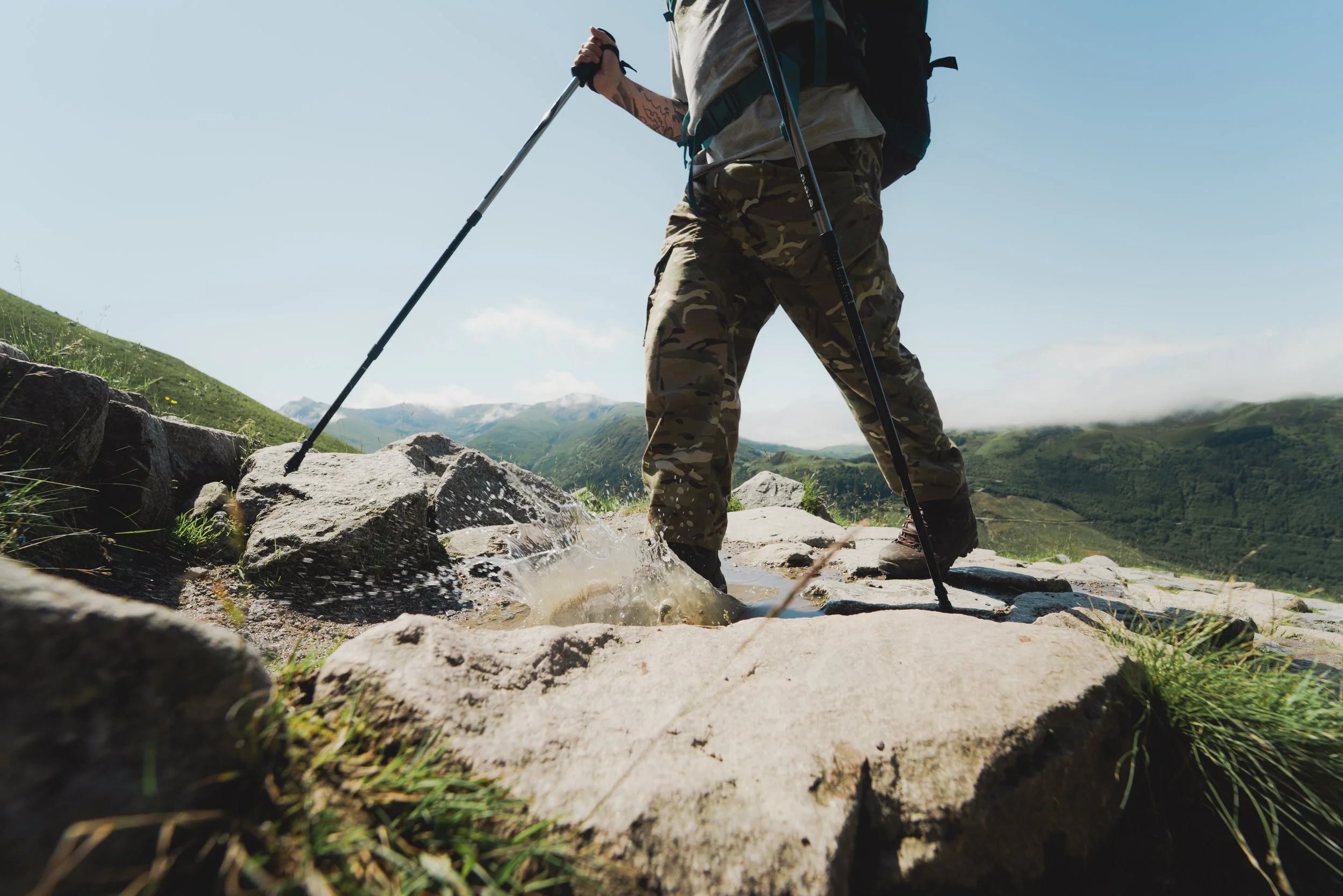 A person hiking in outdoor terrain with a backpack, using trekking poles, crossing a small rocky stream with a backdrop of green hills and a clear blue sky.