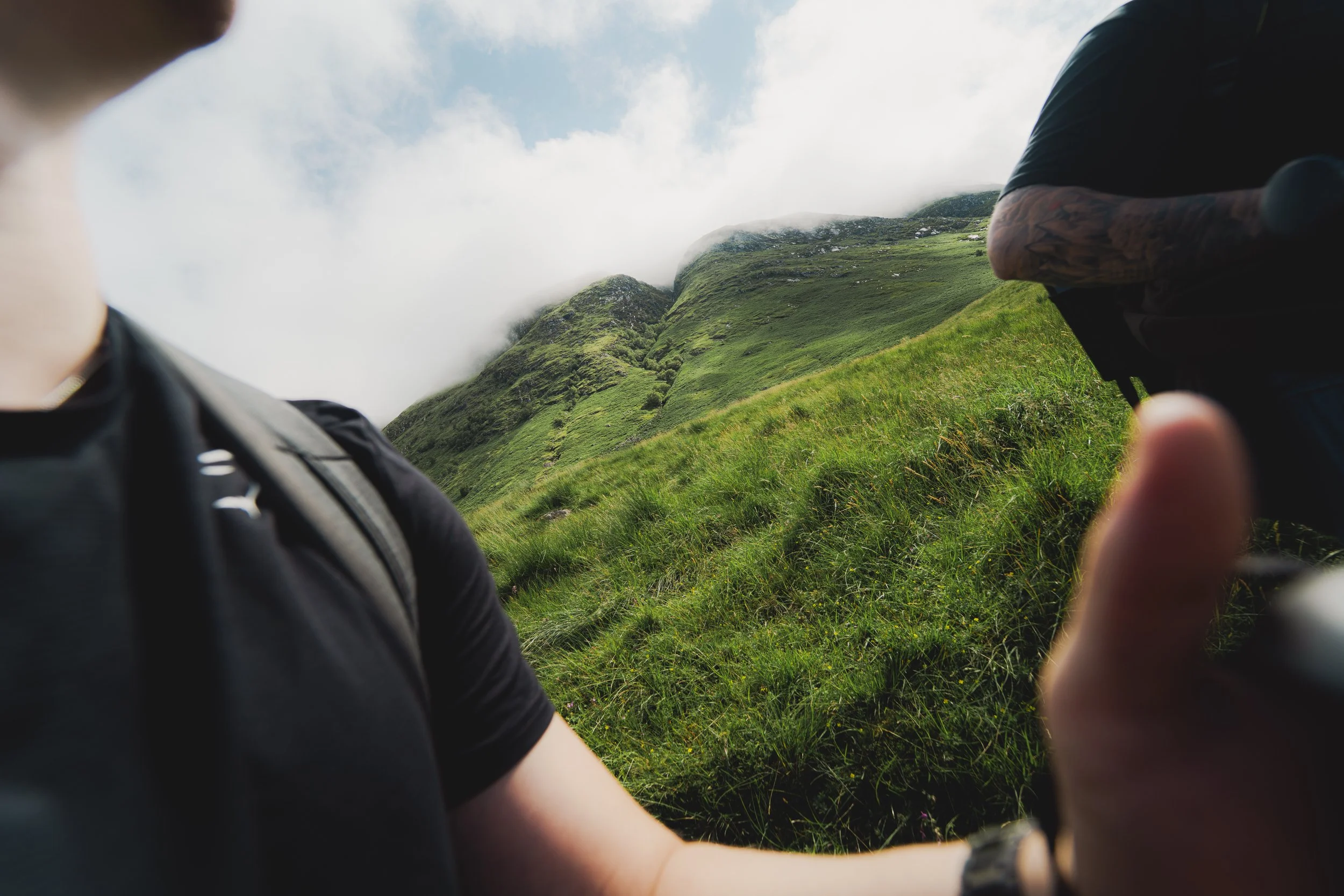 A person wearing a black shirt with a zipper is taking a photo or selfie in a green mountainous area with grass and clouds in the background, with another person with a tattooed arm visible on the right.