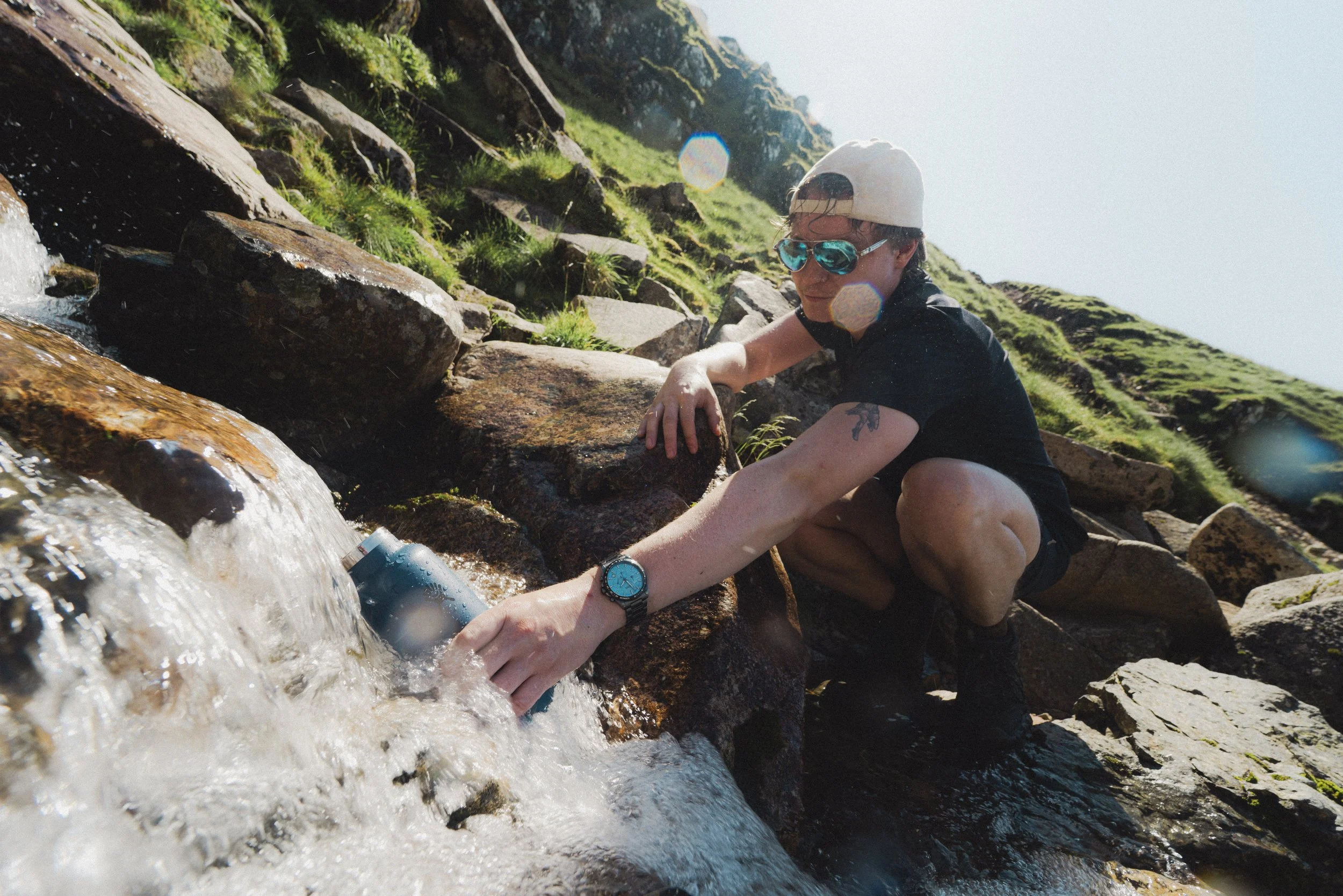Person in sunglasses and a cap reaching into a small stream surrounded by rocks and greenery on a sunny day.