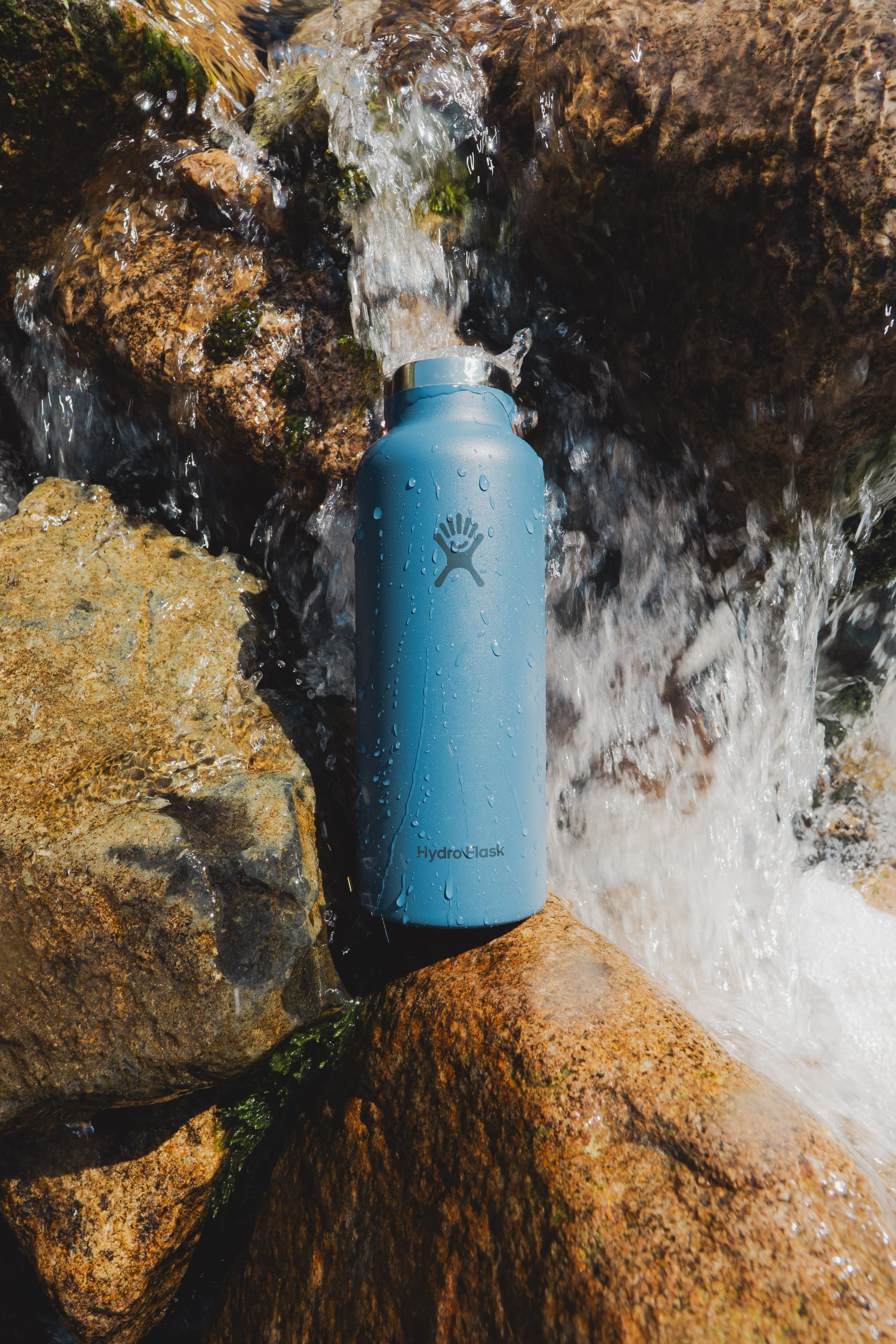 A blue Hydro Flask water bottle placed among rocks by a small waterfall.