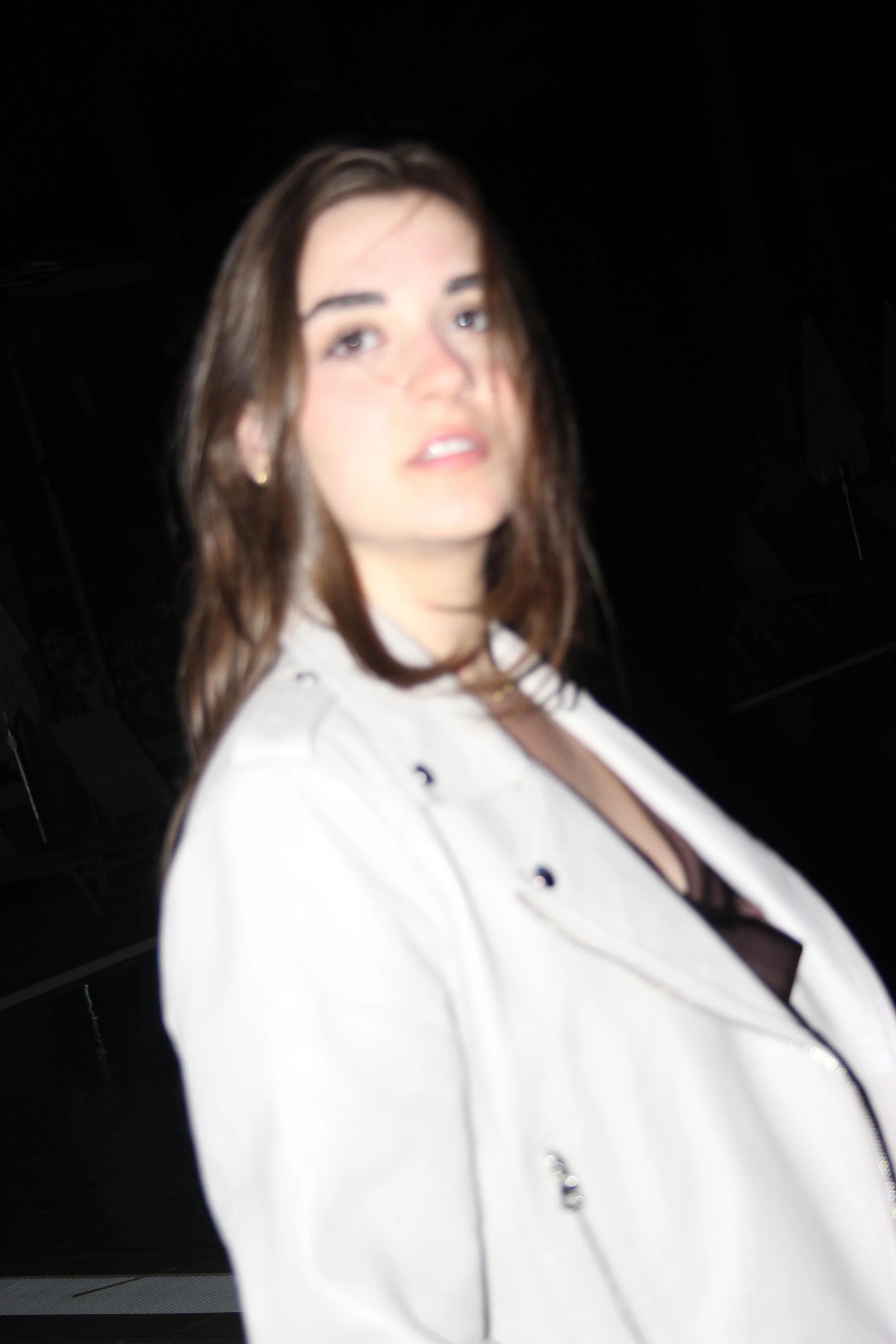 Blurry photo of a young woman with brown hair wearing a white jacket and black top against a dark background.