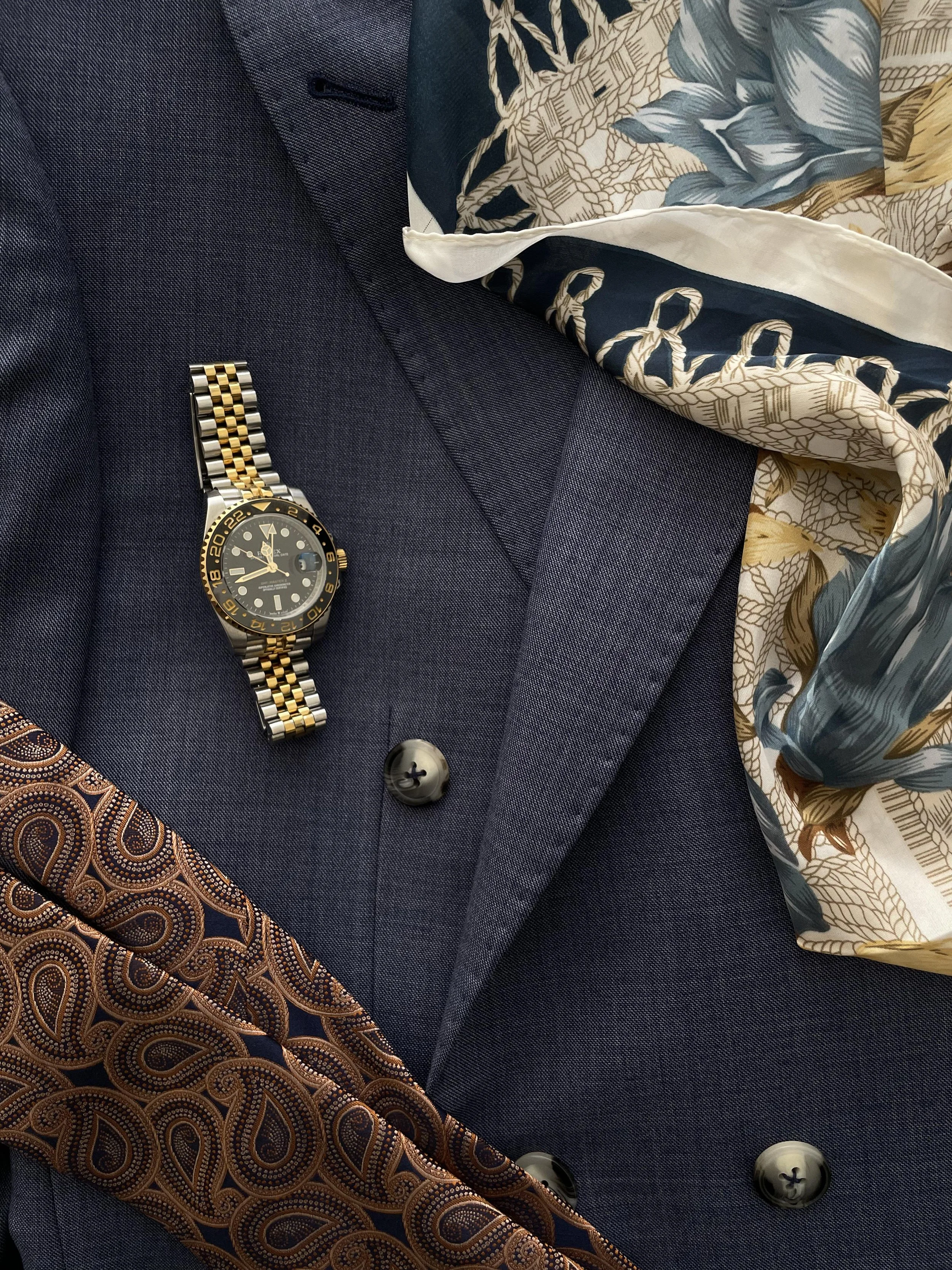 A dark blue suit jacket with two buttons, a black and gold wristwatch, a patterned tie, and a colorful silk scarf.